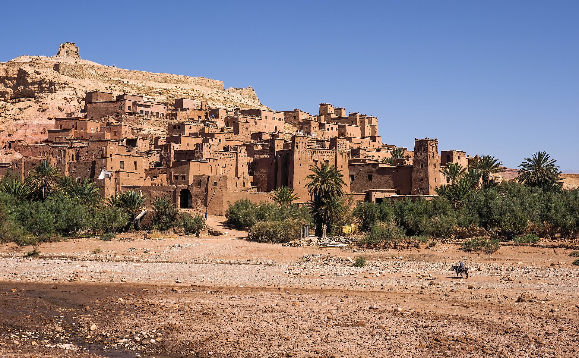 A hillside village of earthen mud-brick houses and towers rises above palm trees and a dry stony riverbed under a deep blue sky