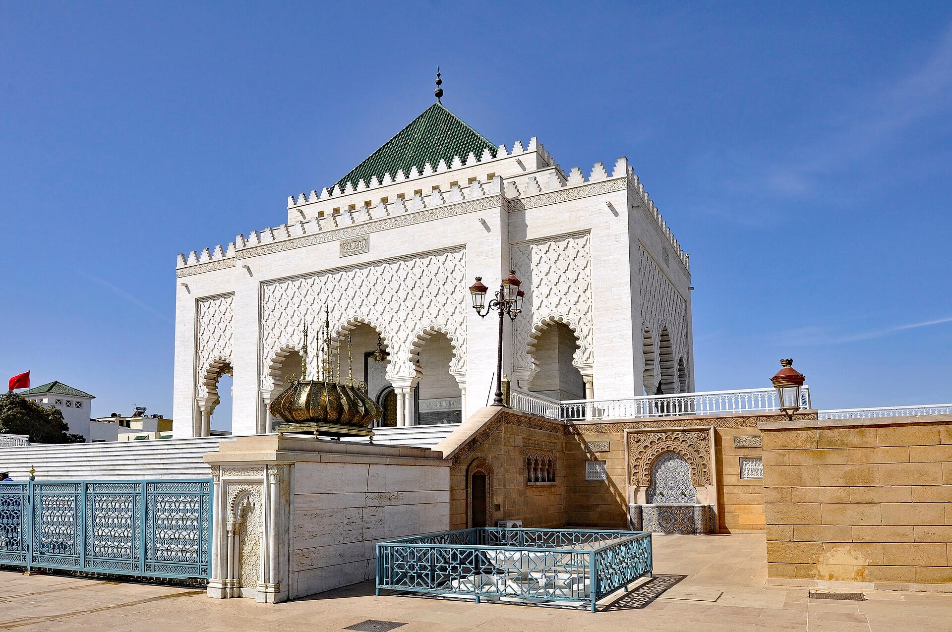 A white marble mausoleum with carved arches and a green tiled pyramid roof stands on a sunny plaza with blue railings and lanterns