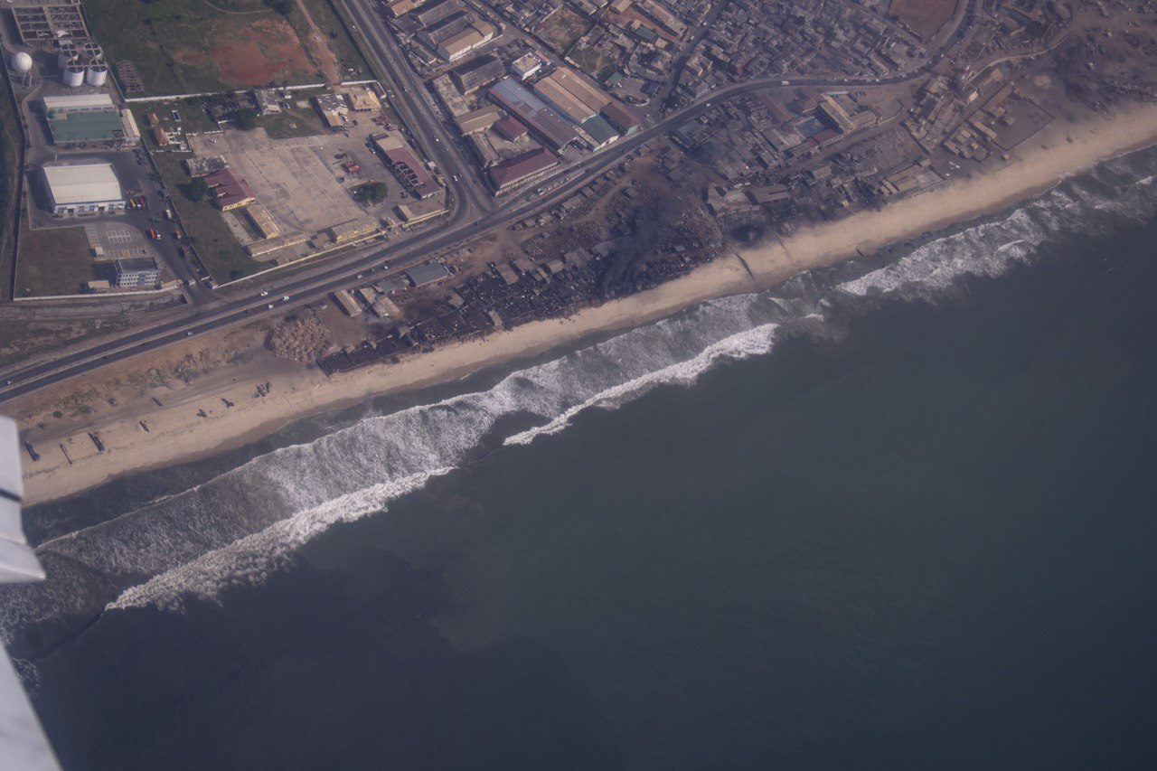 Aerial view of a sandy coastline with breaking waves, a coastal highway and warehouses and houses inland