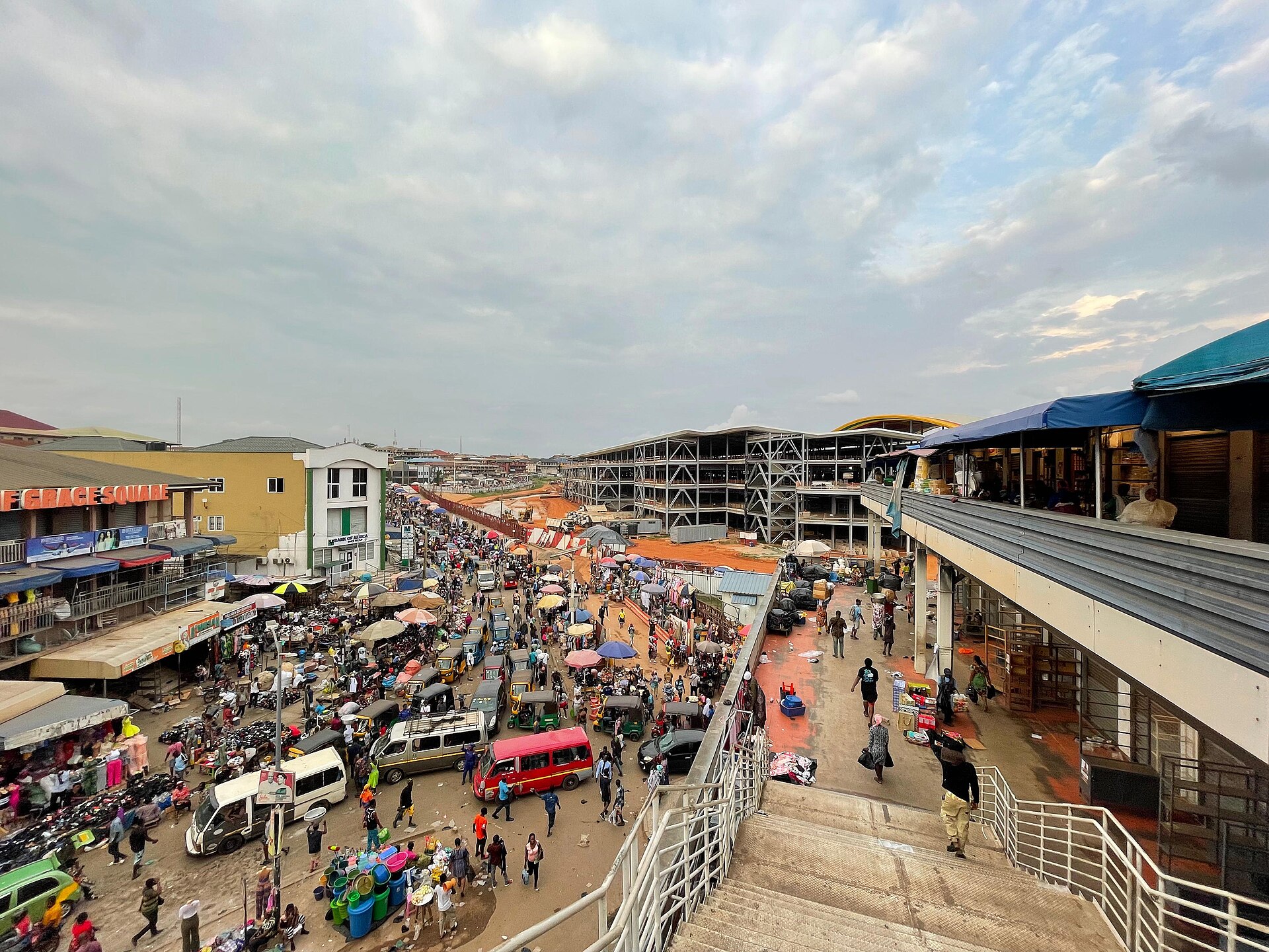 A crowded market street with minibuses, umbrellas and shoppers beside a modern multi-storey market hall under a cloudy sky
