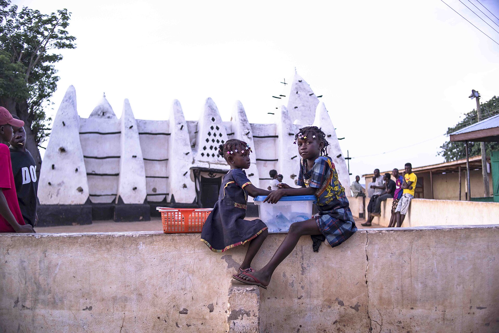 Two girls sit on a low wall in front of a white mud-built mosque with pointed buttresses and wooden beams sticking out