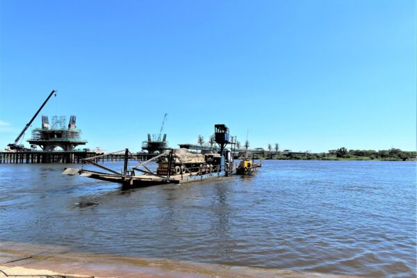 A flat truck ferry carrying a covered lorry sits on the Zambezi River beside a sandy bank, with cranes and bridge piers under construction behind under a clear blue sky