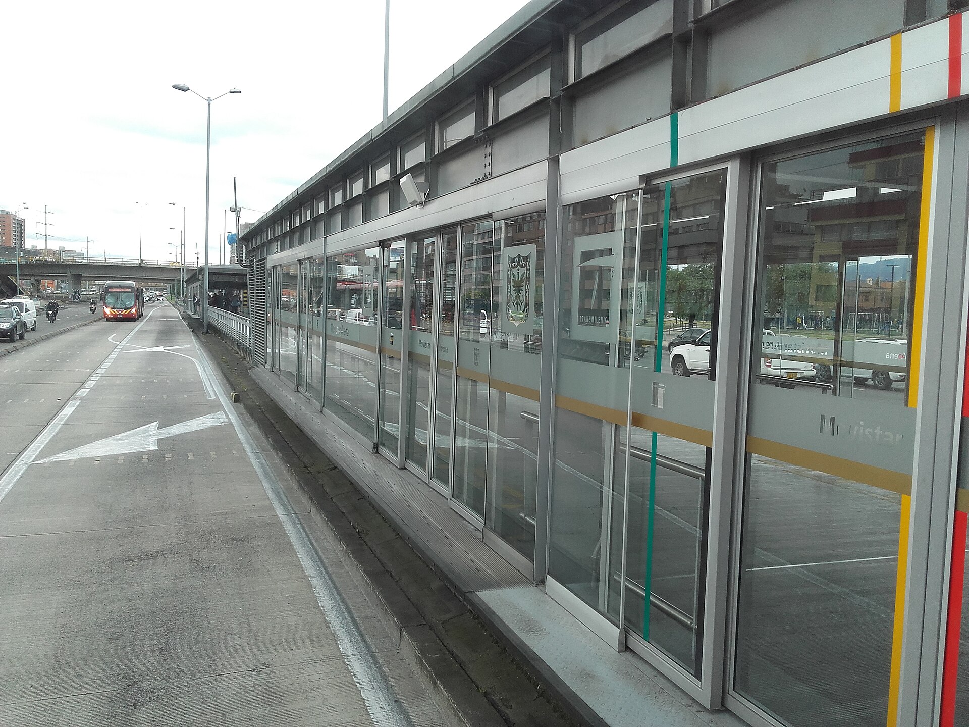Glass doors of the Movistar Arena TransMilenio station on Avenida NQS in Bogotá, with a red bus approaching in the dedicated lane