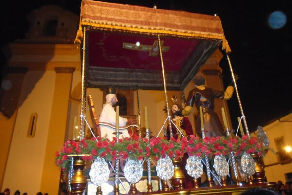A night procession float with painted statues, red flowers and silver ornaments passing a yellow church in Popayán, Colombia.