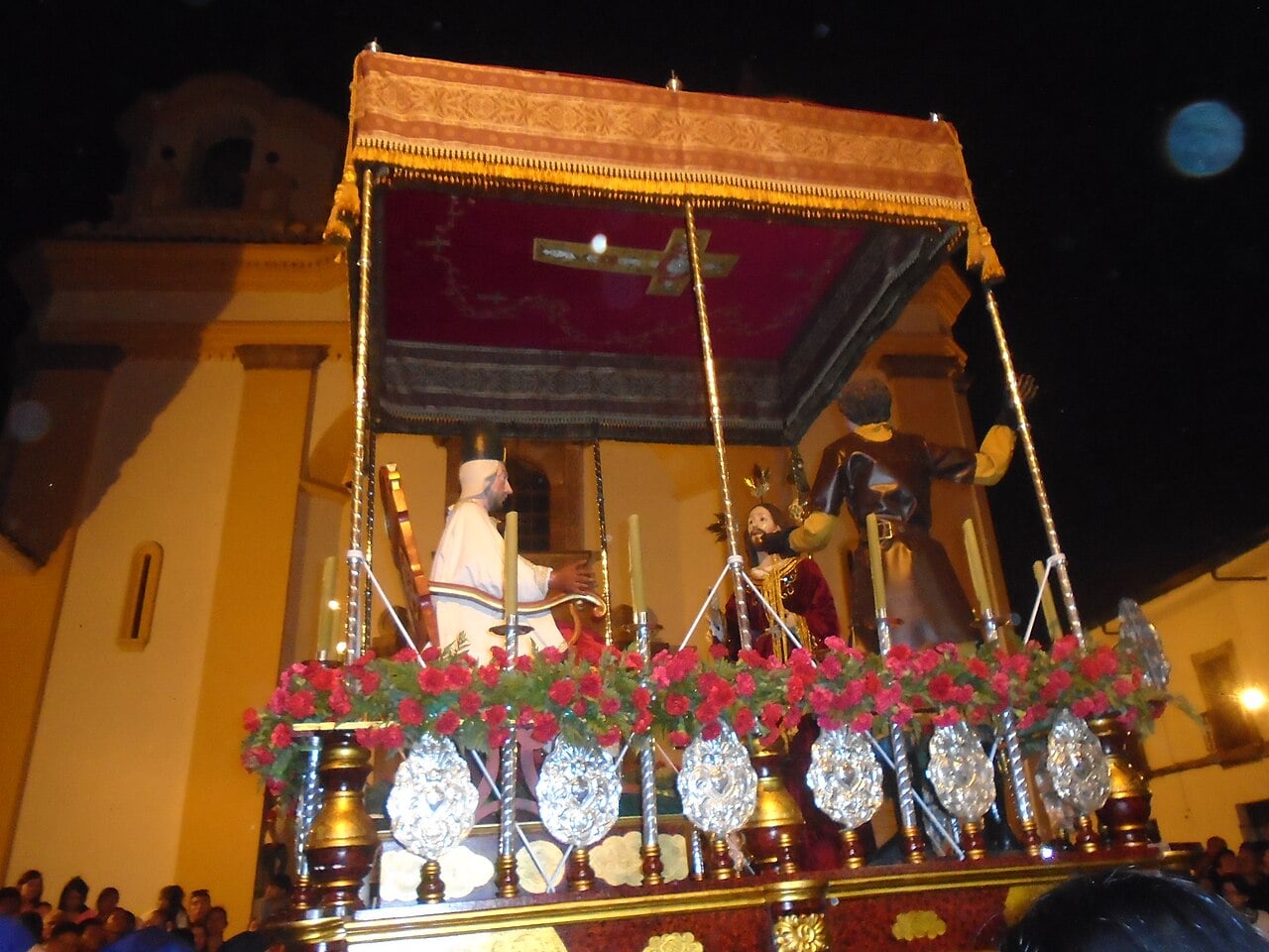 A night procession float with painted statues, red flowers and silver ornaments passing a yellow church in Popayán, Colombia.