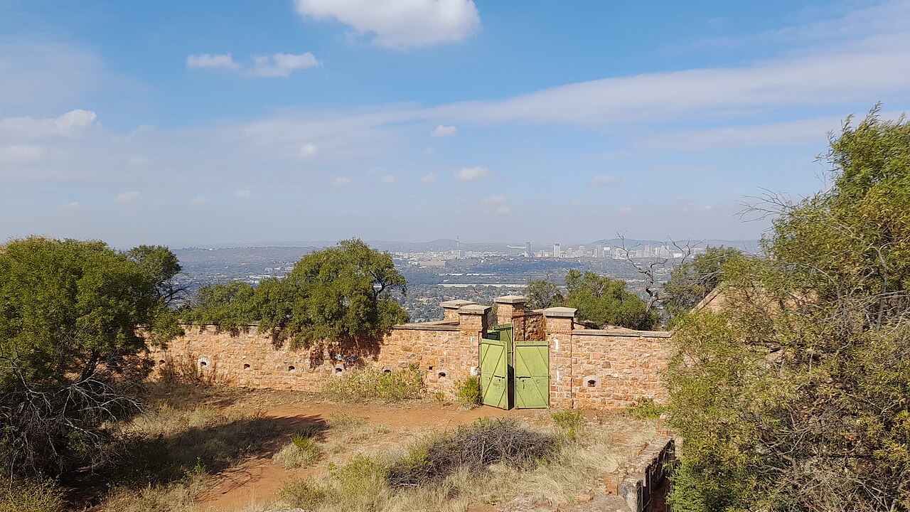 A sandstone fort wall with green gates among trees on a hill, with the Pretoria skyline hazy in the distance under a blue sky.