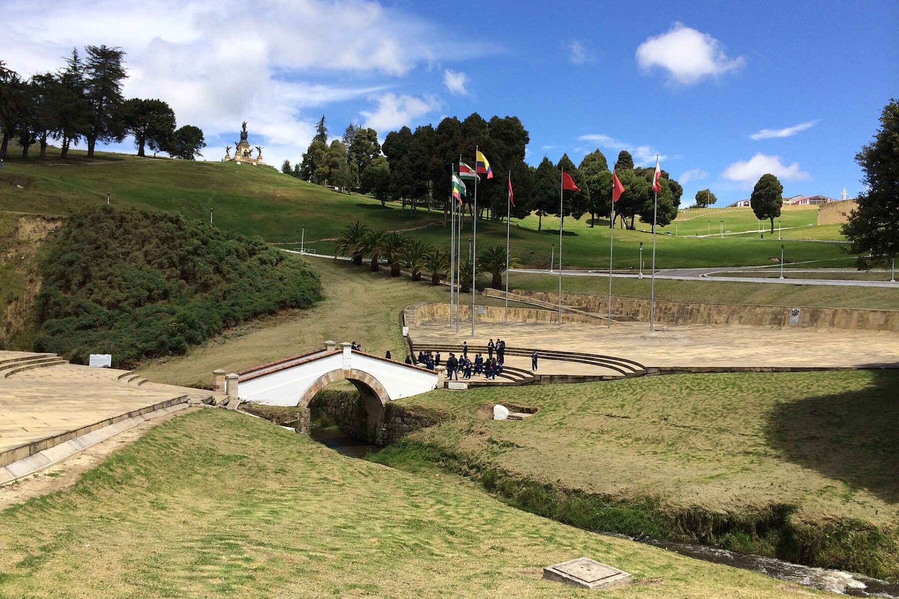 The small white arched Puente de Boyacá bridge over a stream, with a paved plaza, a row of flags and a hilltop monument on green hills under a blue sky