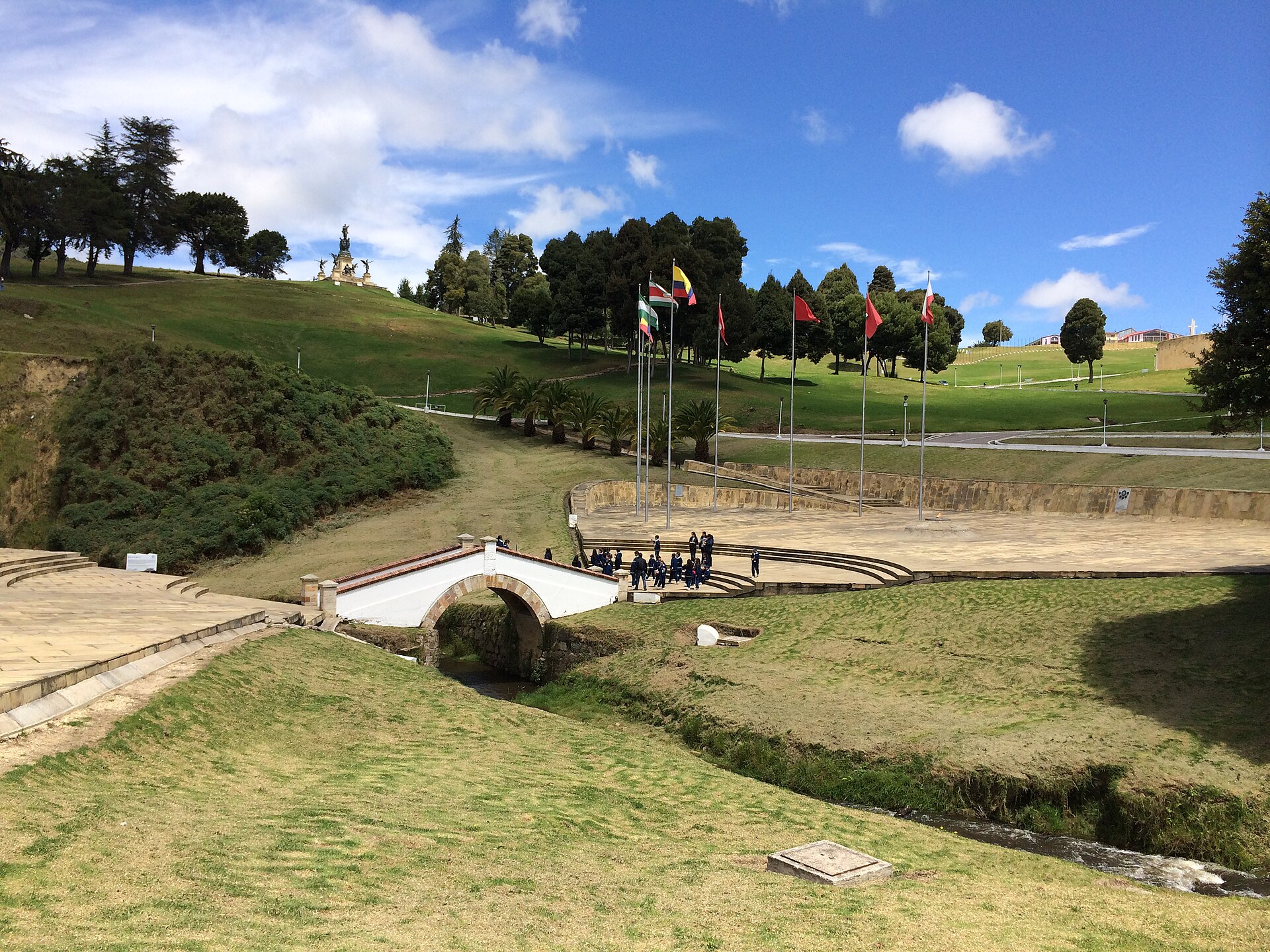 The small white arched Puente de Boyacá bridge over a stream, with a paved plaza, a row of flags and a hilltop monument on green hills under a blue sky