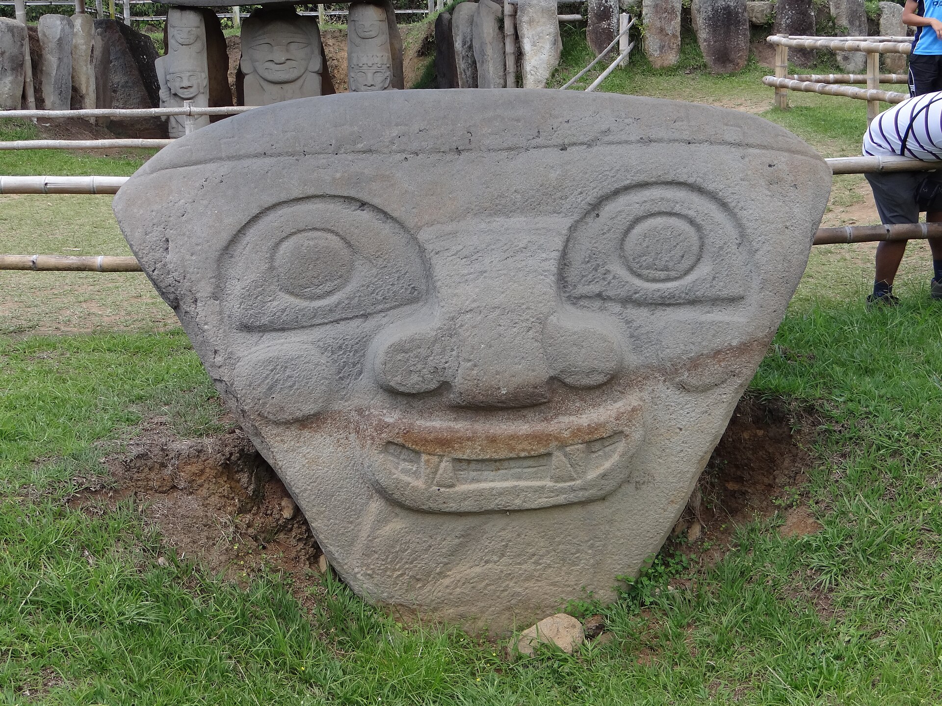 A large triangular carved stone head with round eyes, a broad nose and a fanged grin set in grass, with more stone statues behind a bamboo fence
