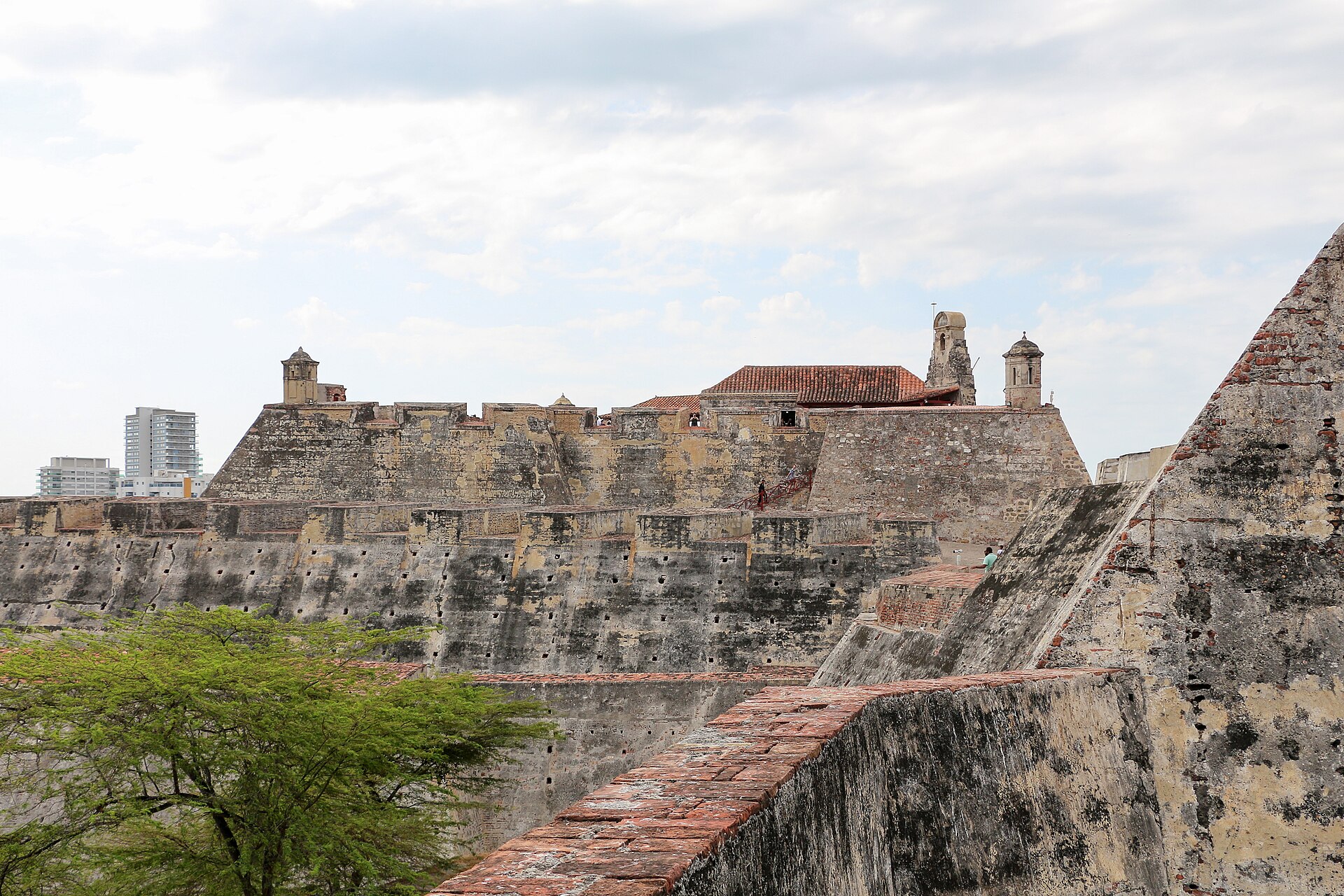 Weathered stone ramparts and sentry towers of Castillo San Felipe de Barajas in Cartagena under a cloudy sky, with a tree in front and modern towers in the distance