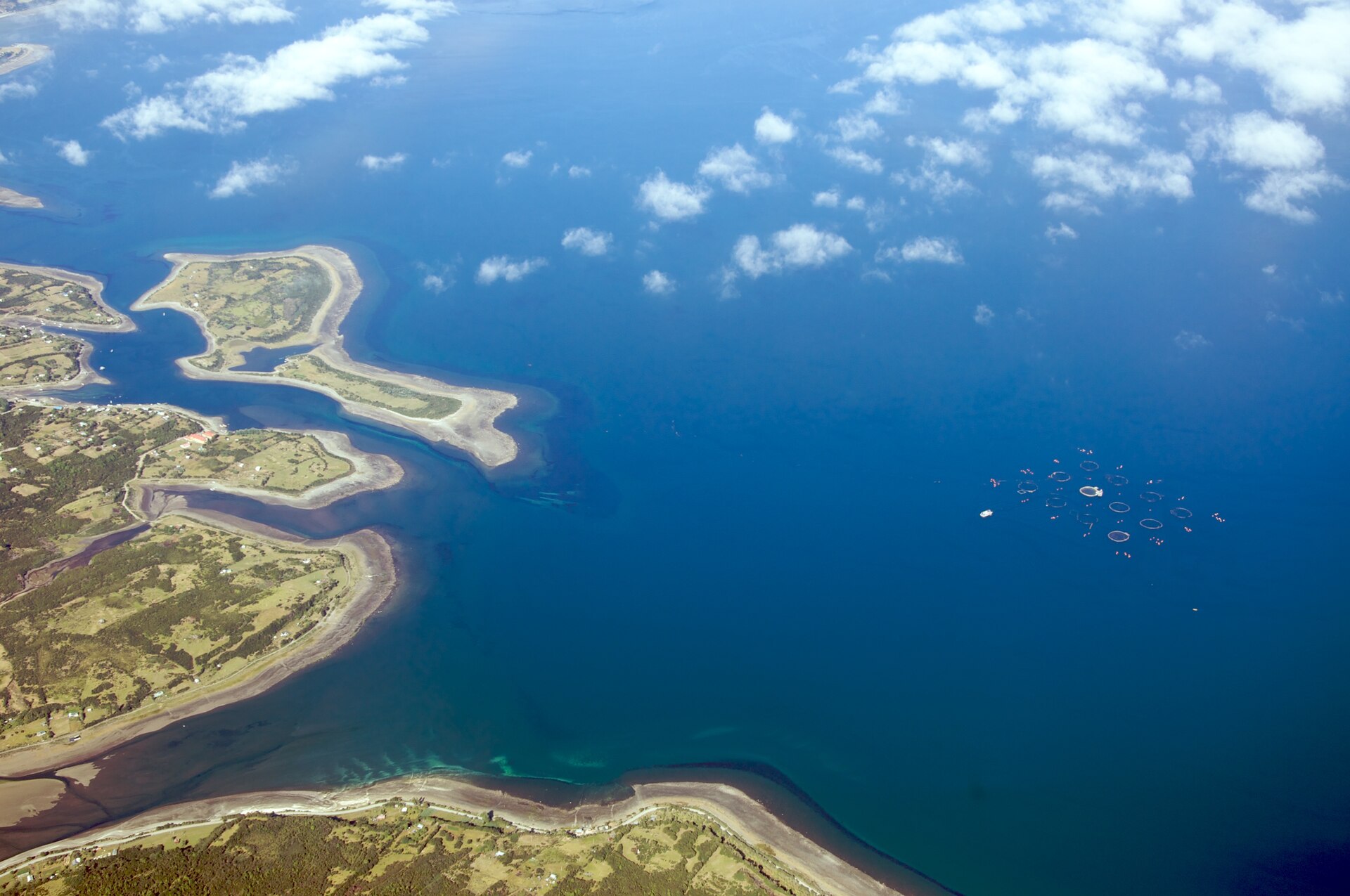 Aerial view of a cluster of circular salmon cages in deep blue water off a green coastline with sandy spits under scattered clouds