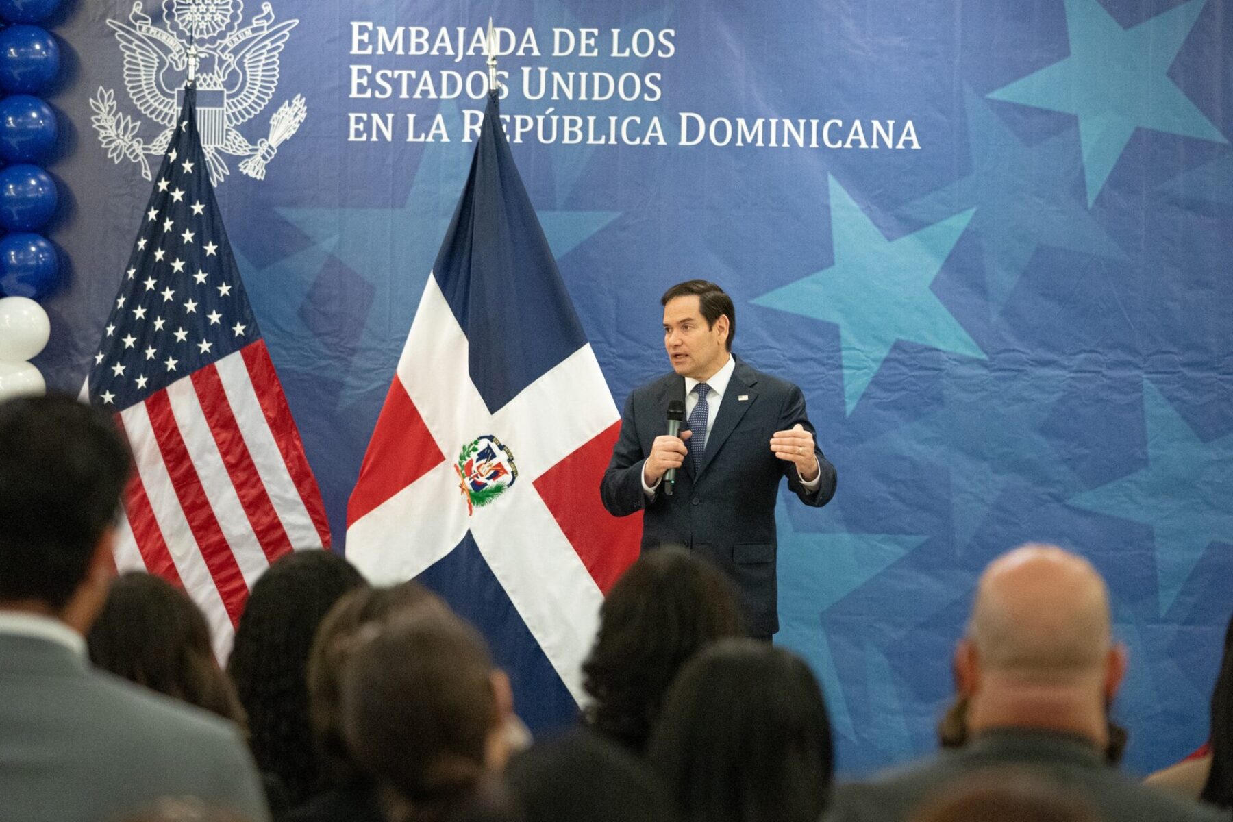 Marco Rubio speaks into a handheld microphone in front of US and Dominican flags and a blue backdrop reading Embajada de los Estados Unidos en la Republica Dominicana, with an audience in the foreground