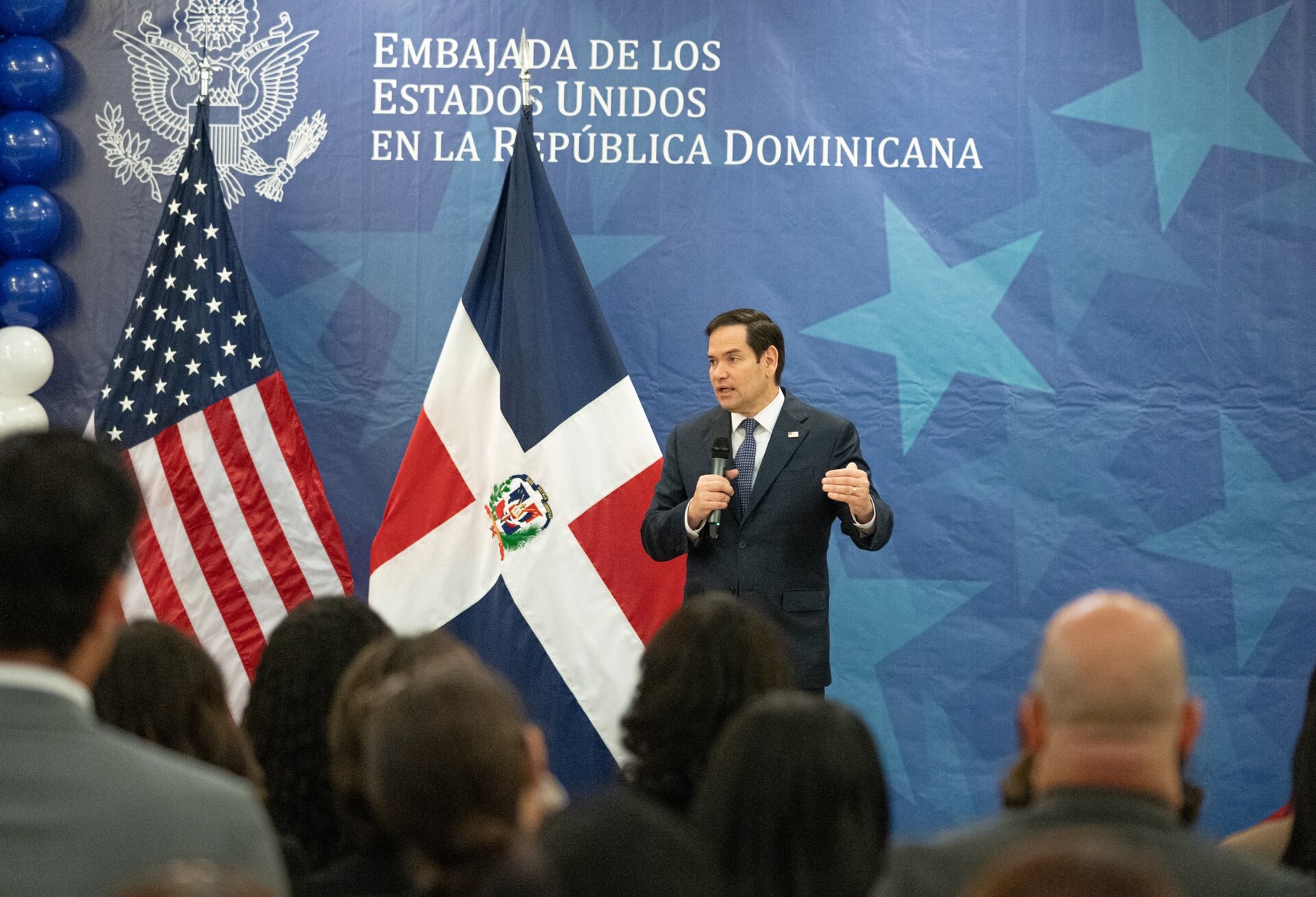 Marco Rubio speaks into a handheld microphone in front of US and Dominican flags and a blue backdrop reading Embajada de los Estados Unidos en la Republica Dominicana, with an audience in the foreground