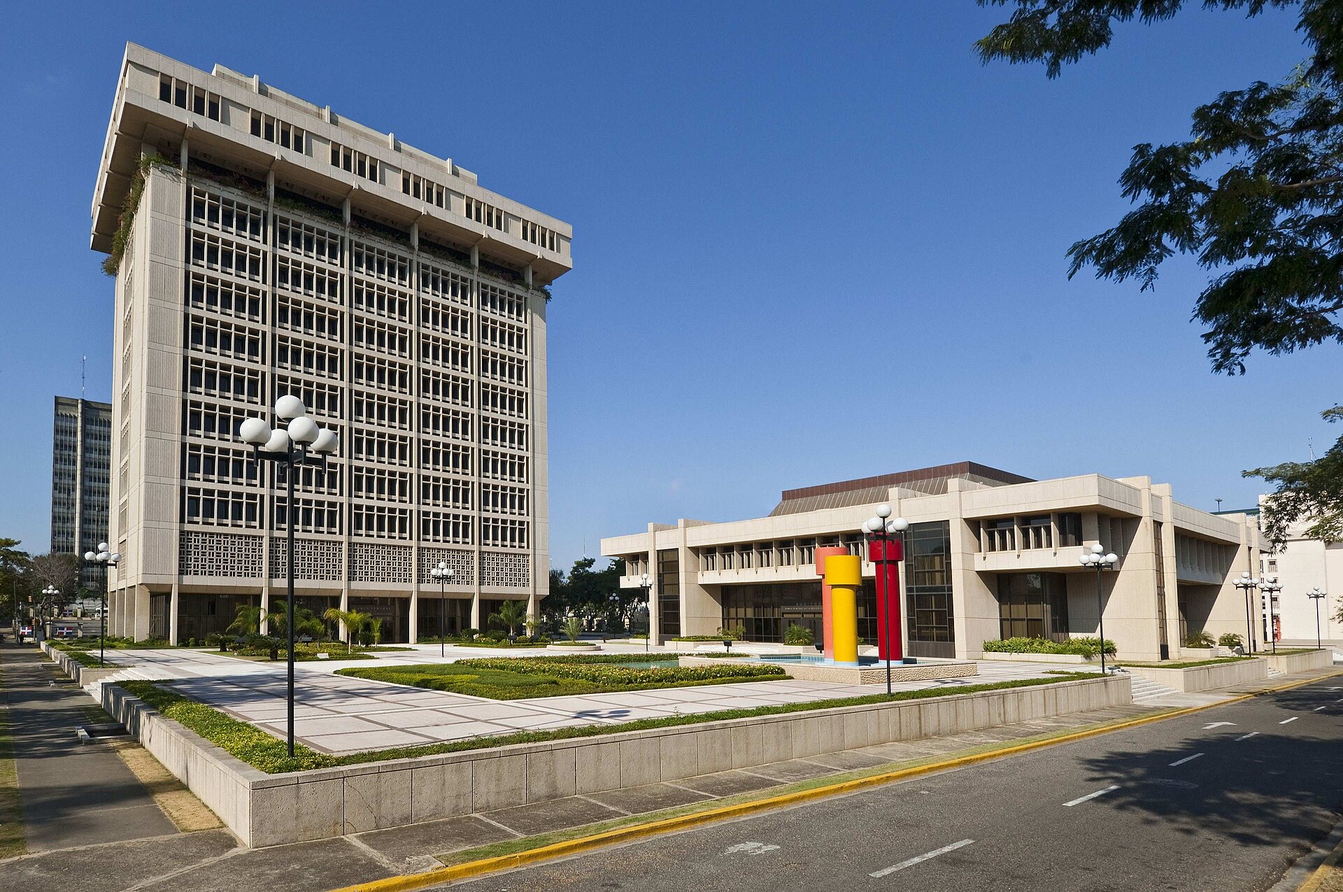A tall concrete office tower and a lower modernist building with a red and yellow sculpture in front, a paved plaza and an empty avenue under a blue sky