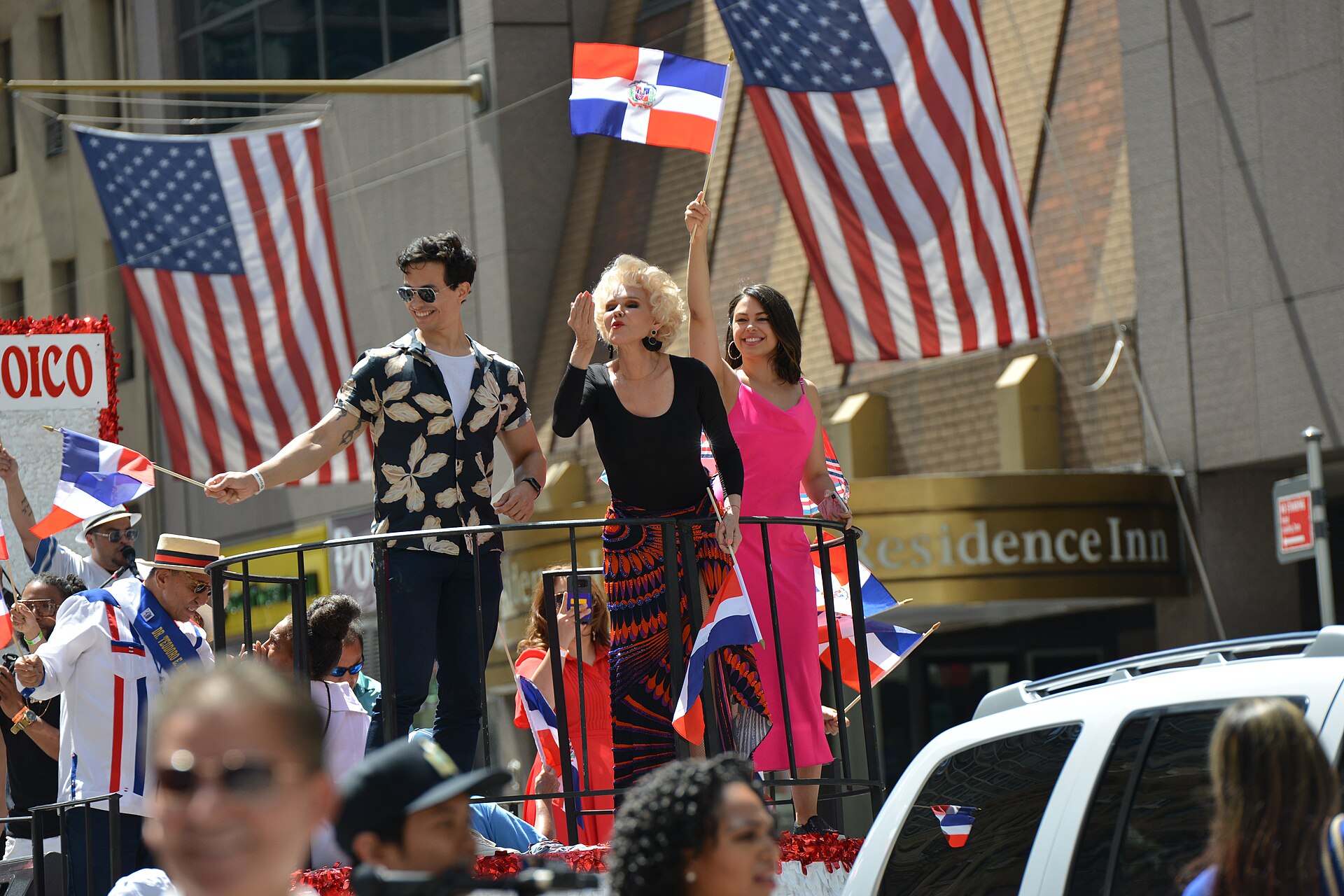 Three performers wave from a parade float decorated with Dominican flags, with large US flags hanging from a building behind them and spectators in front