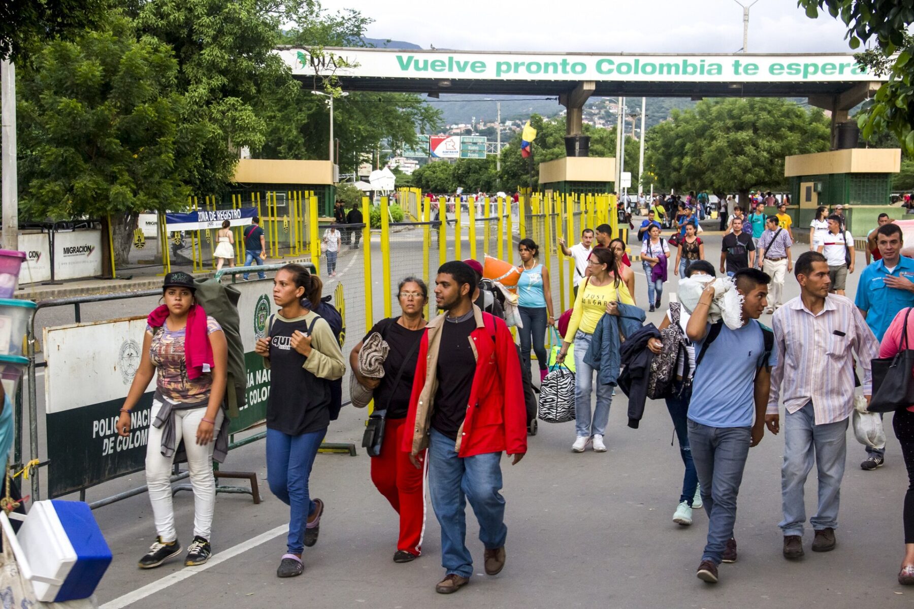 Venezuelans carrying bags, blankets and backpacks walk across the border into Colombia under a sign reading Vuelve pronto Colombia te espera, with Colombian police barriers at the side