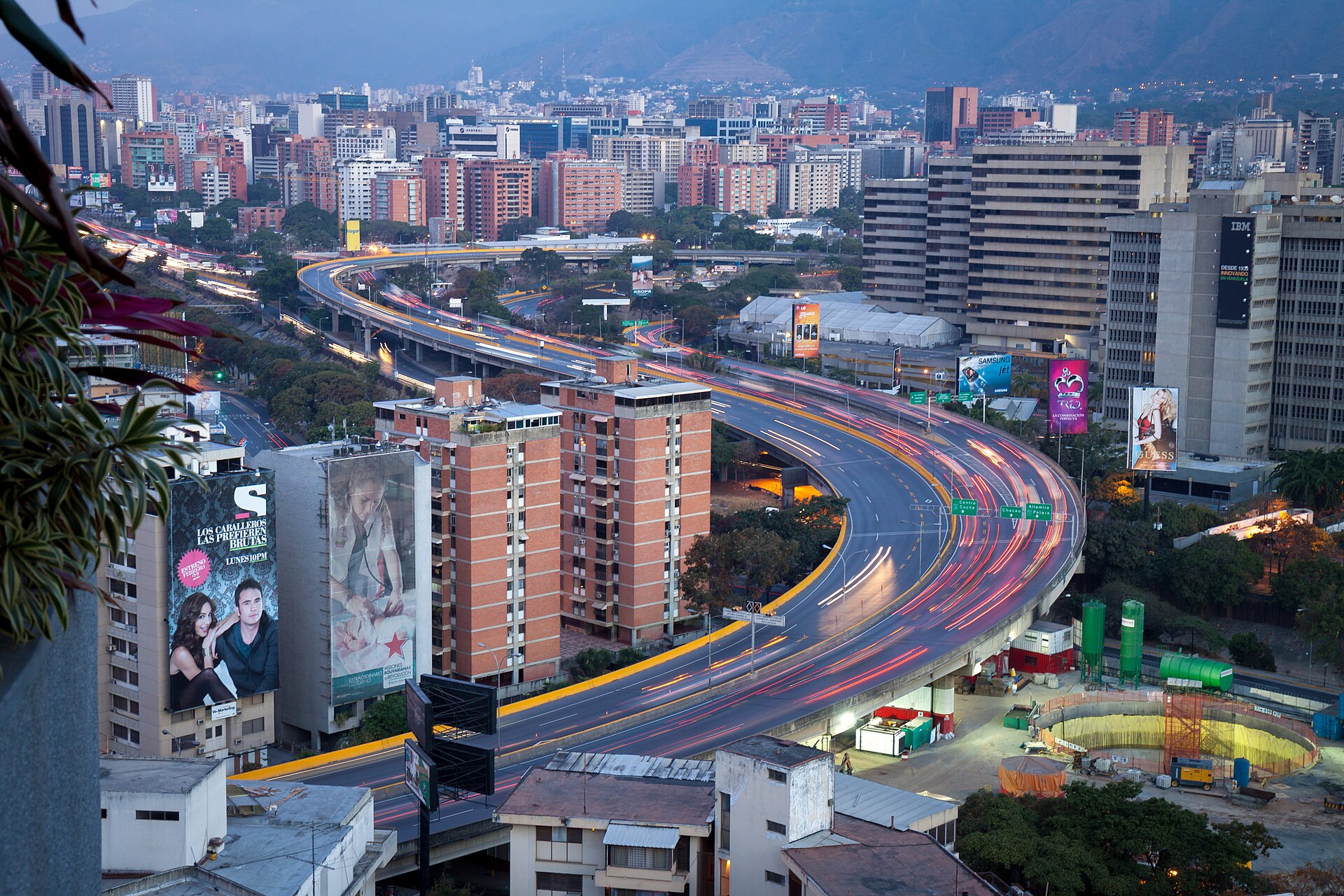 View over Caracas at dawn, with a curving elevated freeway full of light trails from traffic, brick apartment towers, office blocks and billboards, and mountains in the background