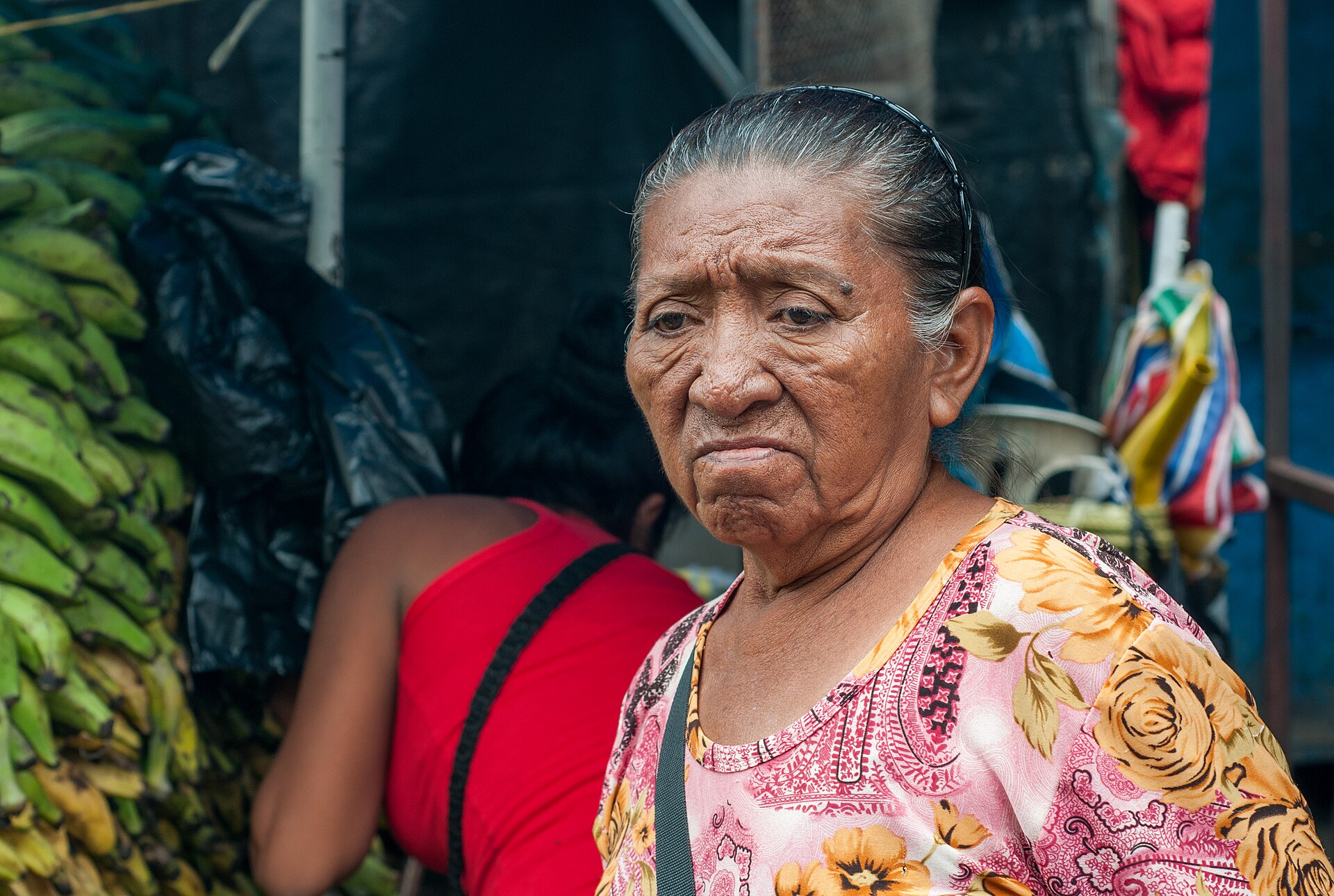 An older Wayuu woman with grey hair pulled back, wearing a pink floral blouse, stands at a market stall with bunches of green plantains and another shopper behind her