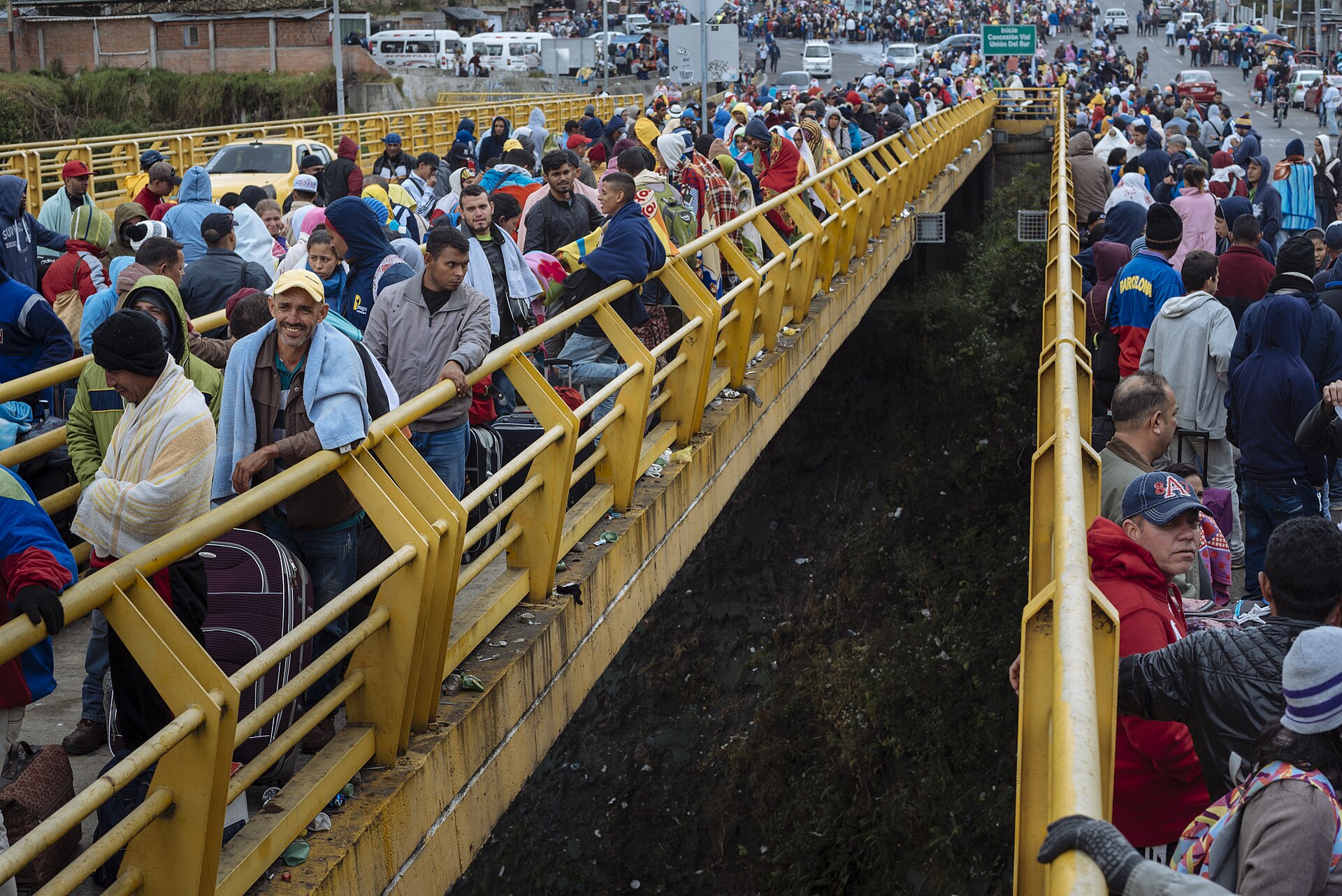 Crowds of Venezuelans wrapped in blankets and carrying suitcases queue along both sides of a bridge with yellow railings, with more people waiting on the road beyond