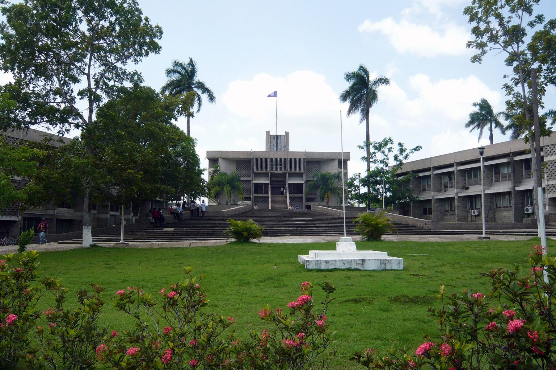 The concrete National Assembly building in Belmopan with the Belize flag on its roof, wide steps, palm trees and a lawn with flowering shrubs in front