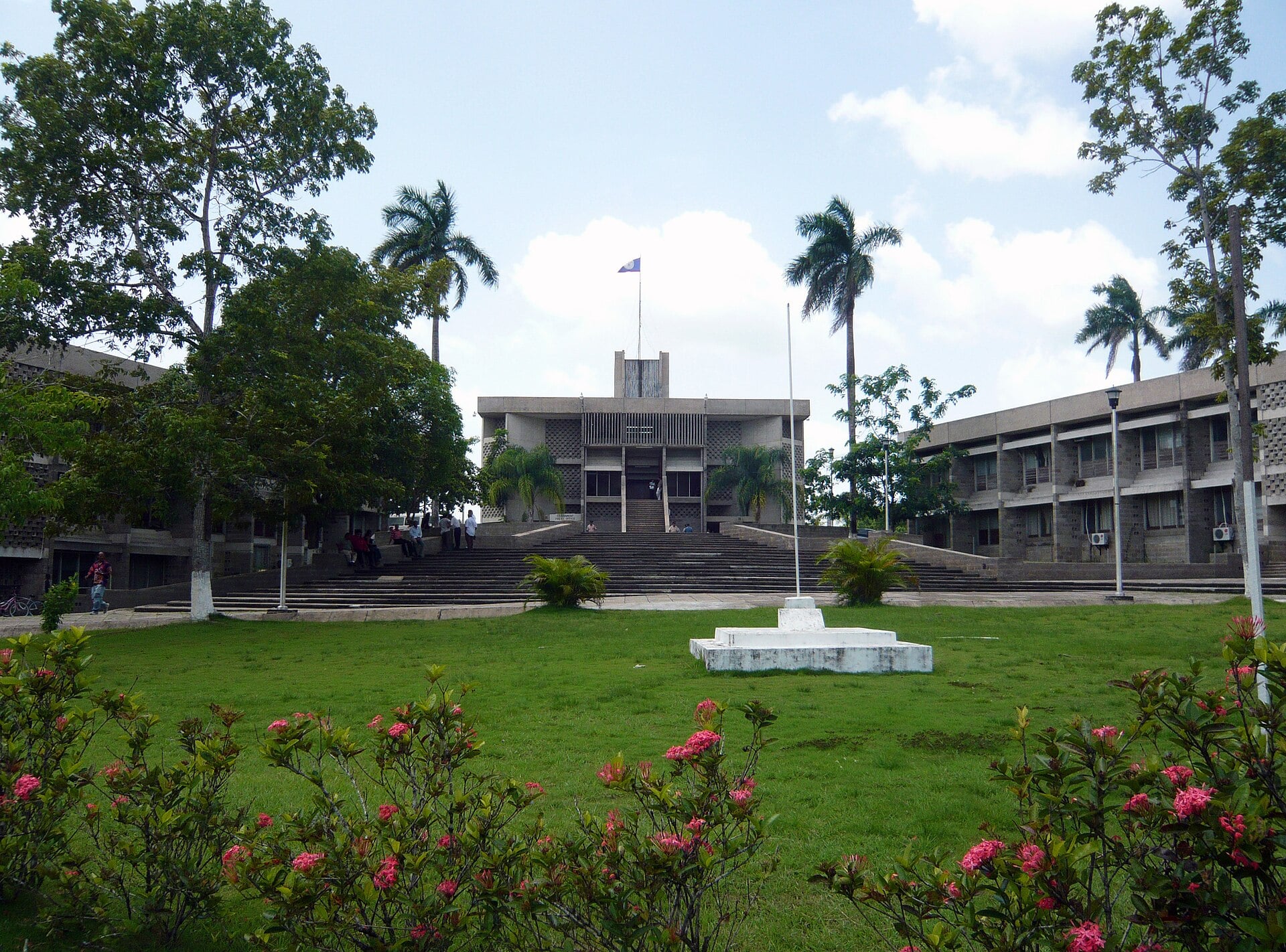 The concrete National Assembly building in Belmopan with the Belize flag on its roof, wide steps, palm trees and a lawn with flowering shrubs in front