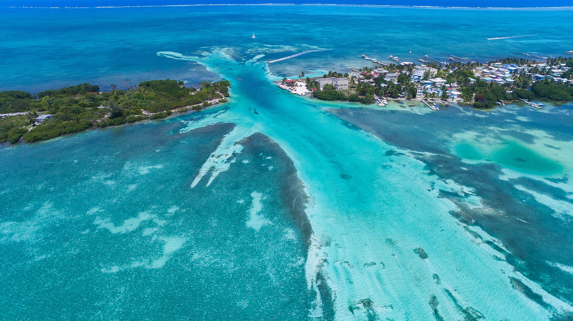 Aerial view of turquoise shallow water around Caye Caulker, with a channel cutting between two wooded parts of the island, houses and piers on the right and a line of surf on the horizon