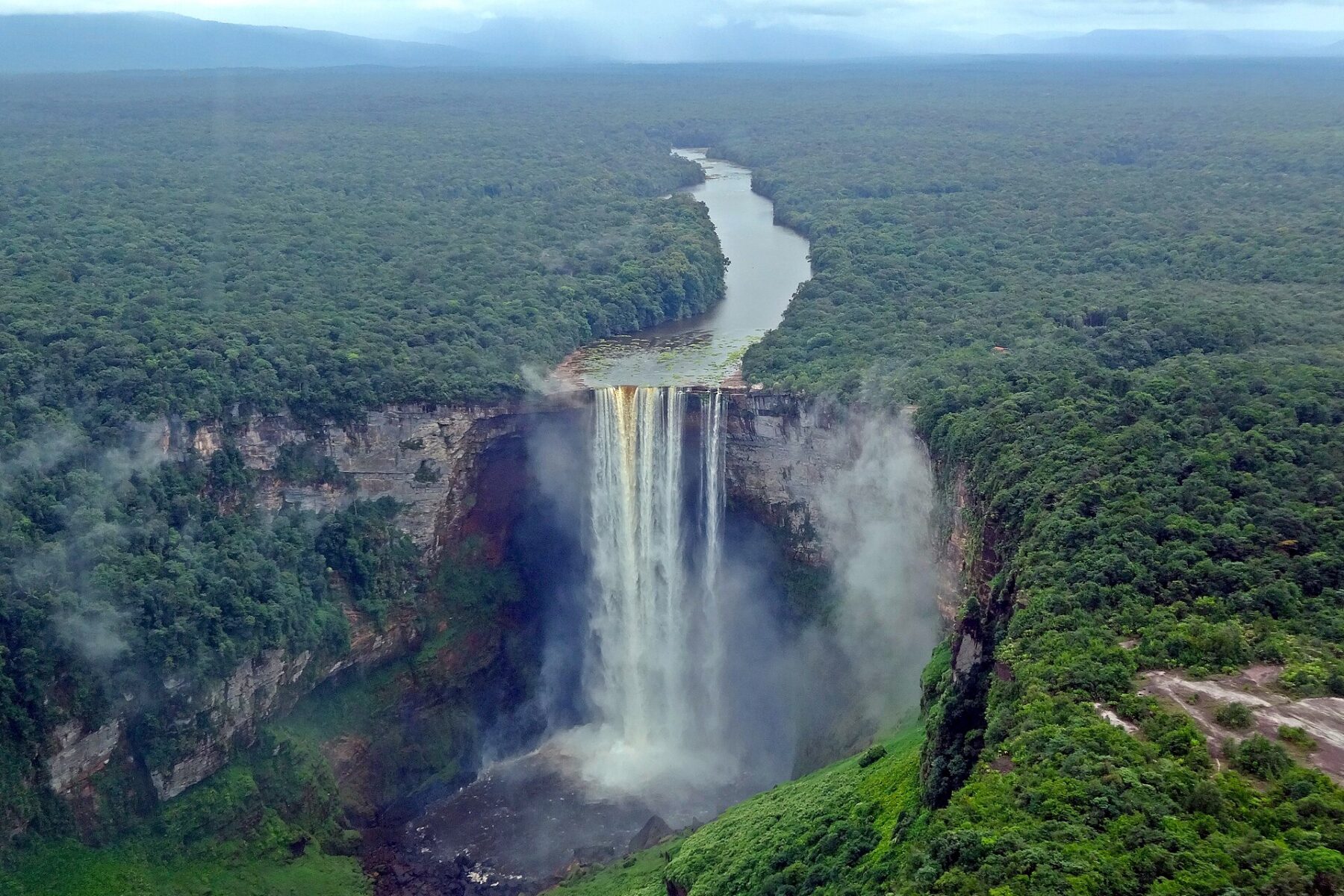 Aerial view of a tall waterfall plunging from a rocky plateau into a misty gorge, with a river winding back through dense green rainforest under low clouds