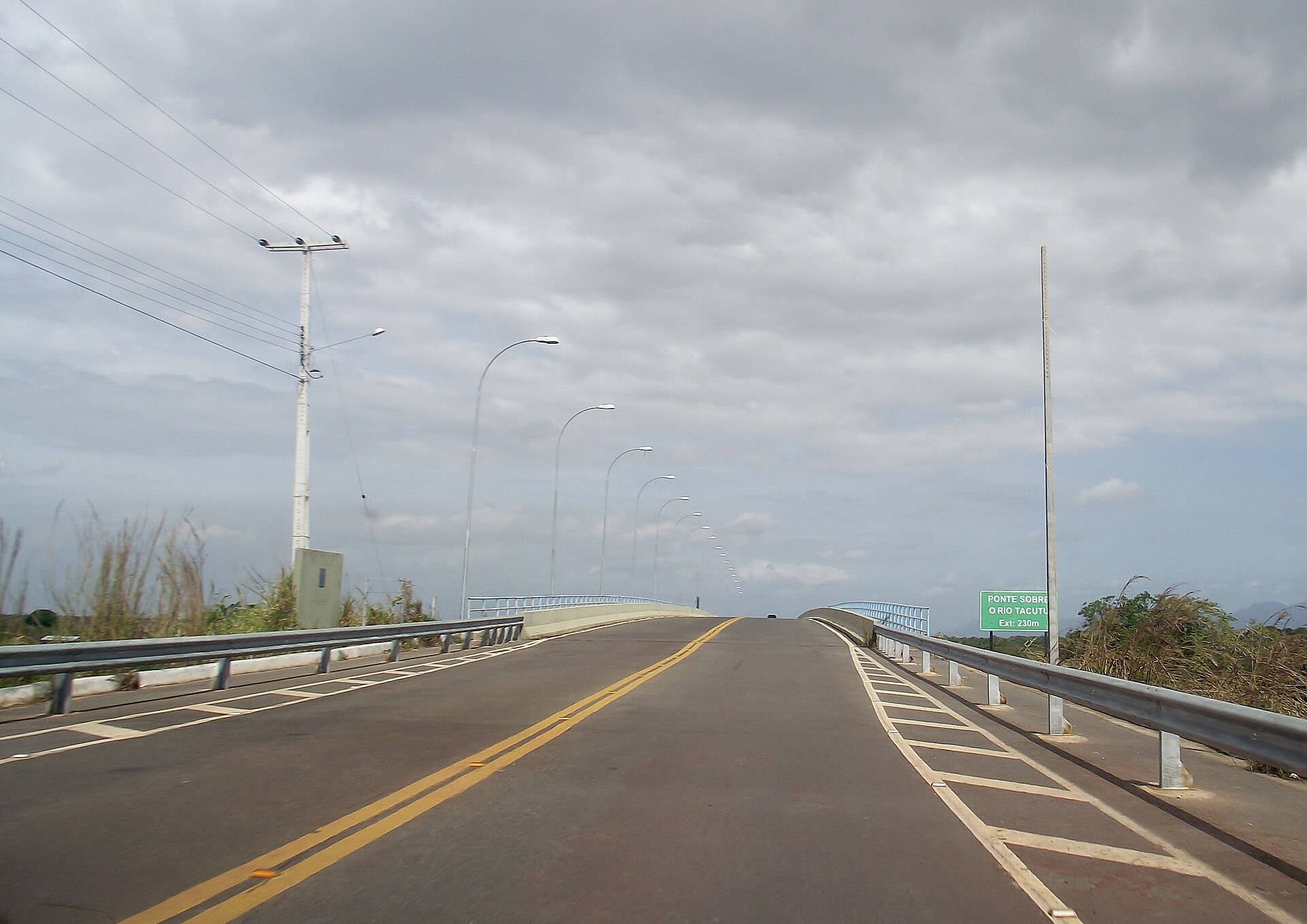 A two-lane road with yellow centre lines and street lamps rising onto a bridge under a cloudy sky, with a green sign in Portuguese naming the Tacutu River