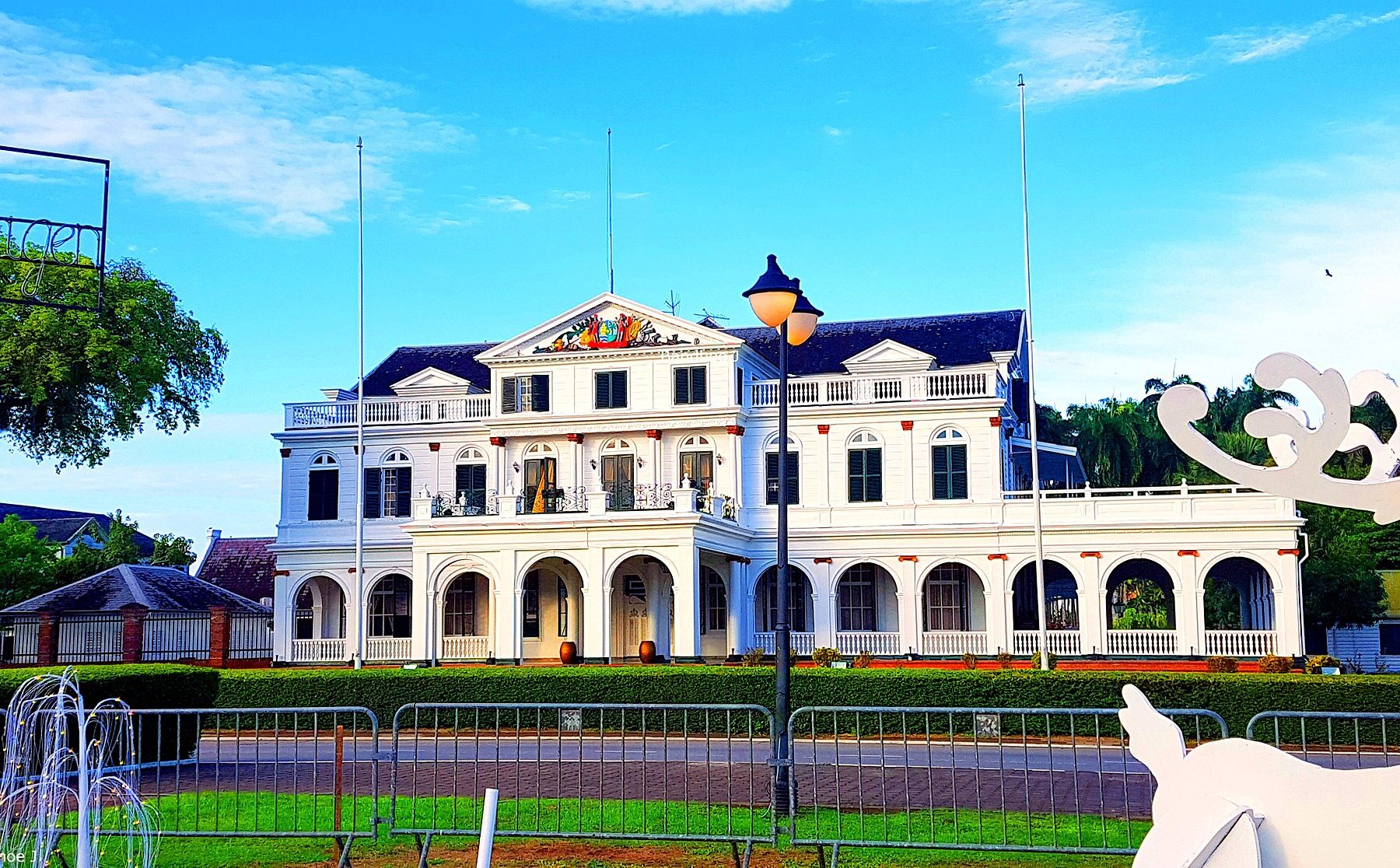 The white two-storey Presidential Palace of Suriname with arched verandas, balconies and a coat of arms on its central gable, seen across a lawn and a low railing, with a small fountain and a white decorative figure in the foreground under a blue sky