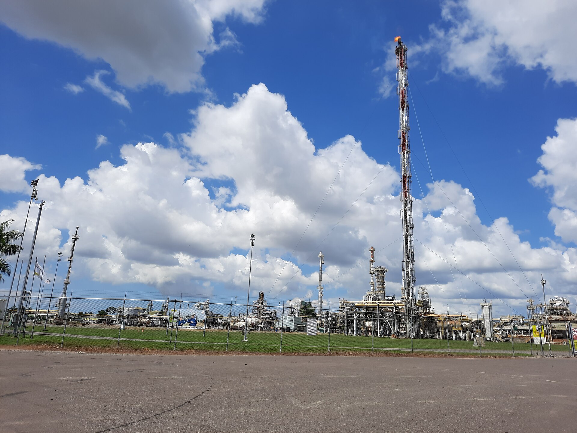 A tall red and white flare stack and the pipes and towers of an oil processing plant behind a fence and a strip of lawn, under a blue sky with large white clouds