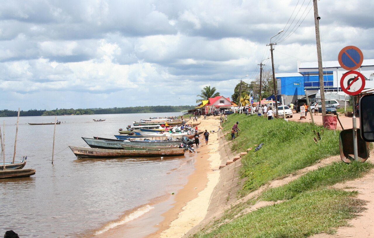 Long wooden river boats pulled up along a sandy bank of a wide brown river, with people on the shore, a grassy embankment, shops, a blue building and traffic signs on the right under a cloudy sky