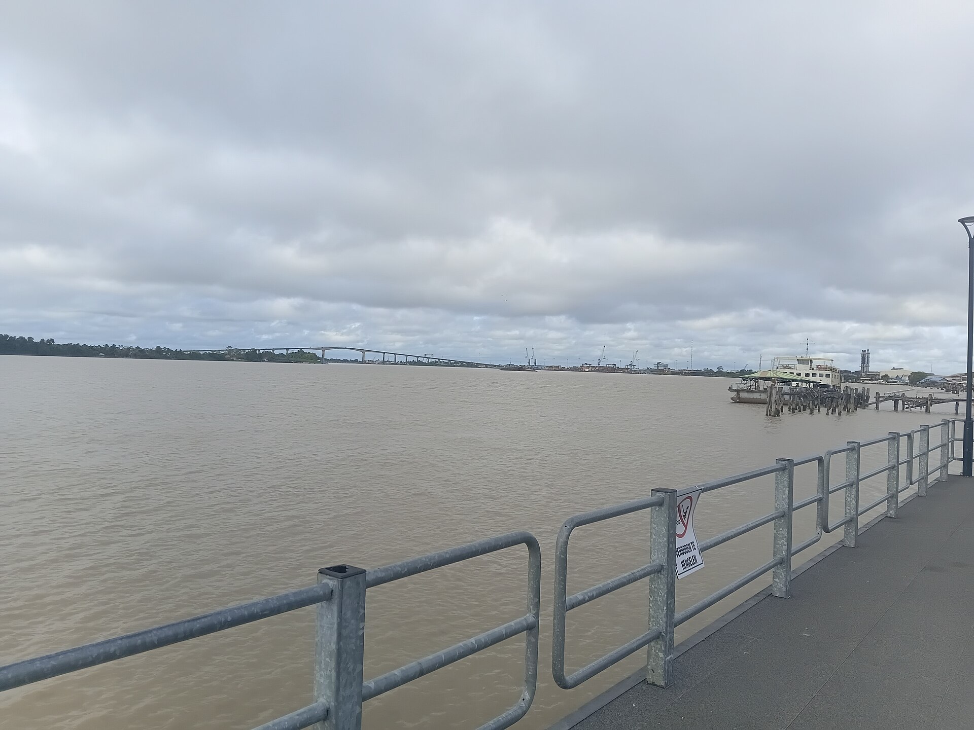 A wide brown river under a grey cloudy sky seen from a railed waterfront promenade, with a long bridge on the horizon and a ferry at a jetty on the right