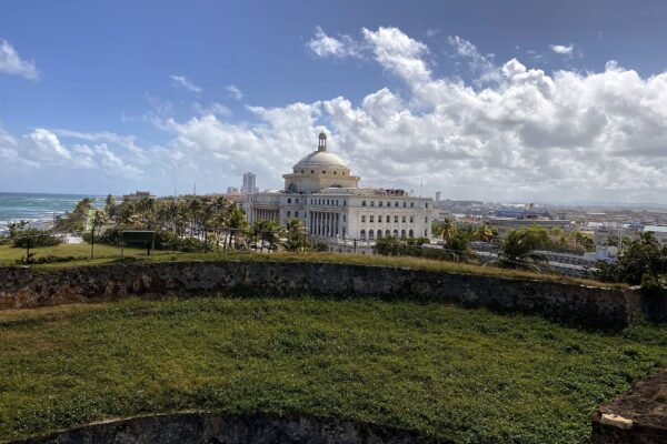 The white domed Capitol of Puerto Rico rises behind palm trees and the grassy stone walls of San Cristóbal Fortress, with the Atlantic Ocean to the left under a blue sky with clouds