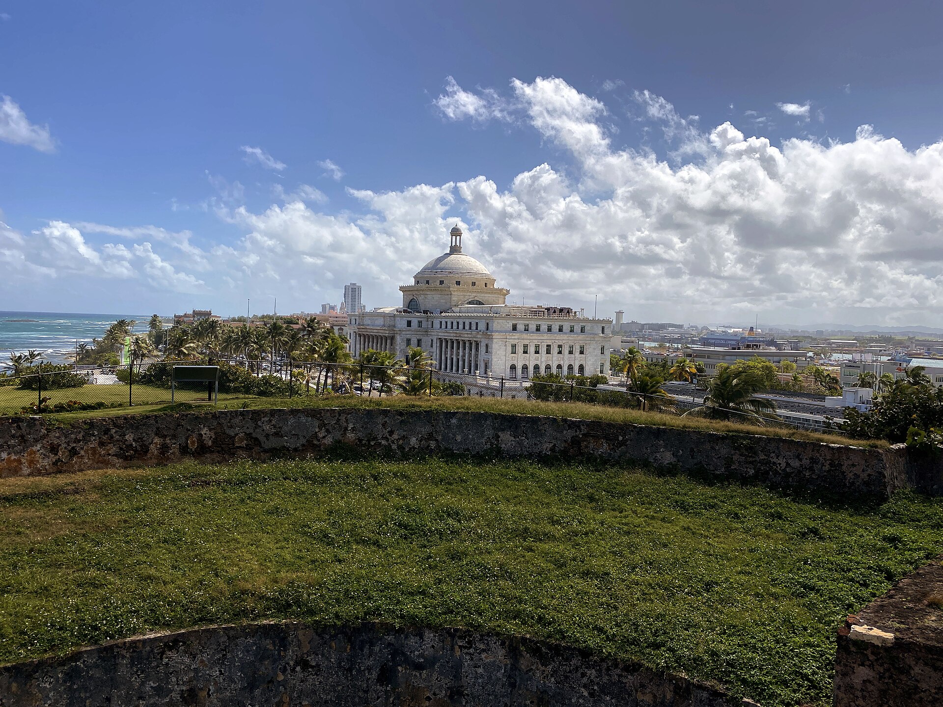 The white domed Capitol of Puerto Rico rises behind palm trees and the grassy stone walls of San Cristóbal Fortress, with the Atlantic Ocean to the left under a blue sky with clouds