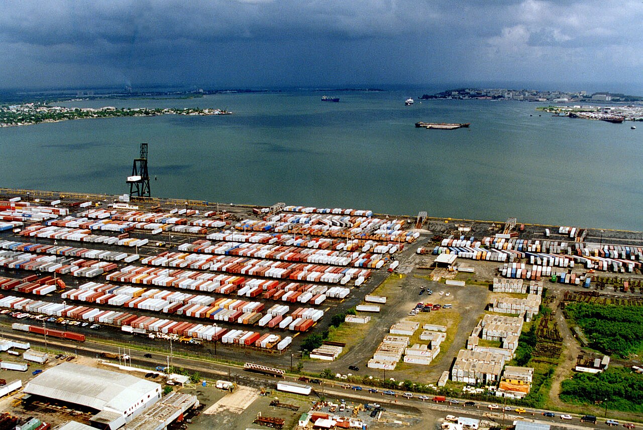 Aerial view of rows of shipping containers stacked in port yards beside San Juan Bay, with ships on the water and the city on the far shore under dark clouds