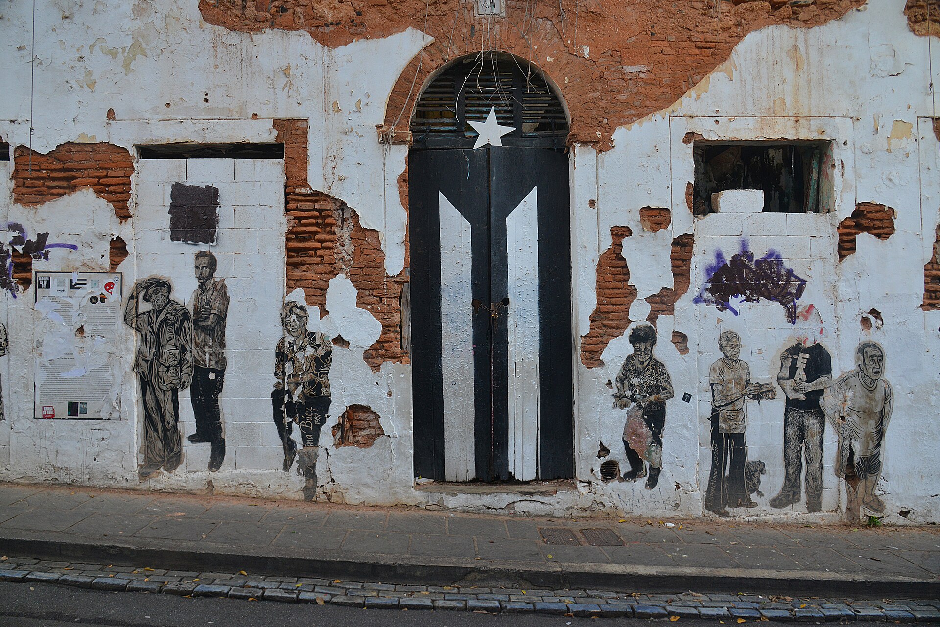 An arched wooden door painted as a black and white Puerto Rican flag with a white star, set in a weathered white wall with exposed brick and pasted-up figures of people