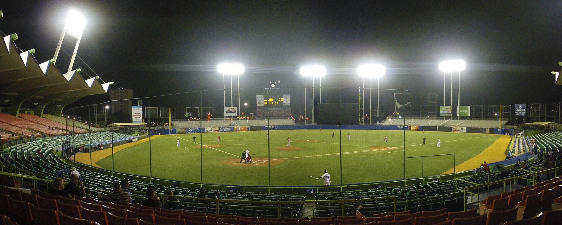 Panoramic view of a floodlit baseball diamond at night inside Hiram Bithorn Stadium, with a batter at the plate and rows of green and red seats in the foreground