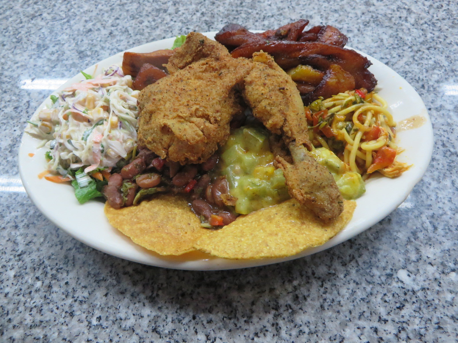 A white plate with fried chicken, red beans, chayote hash, spaghetti, coleslaw, fried ripe plantains and a crisp corn tortilla on a granite counter