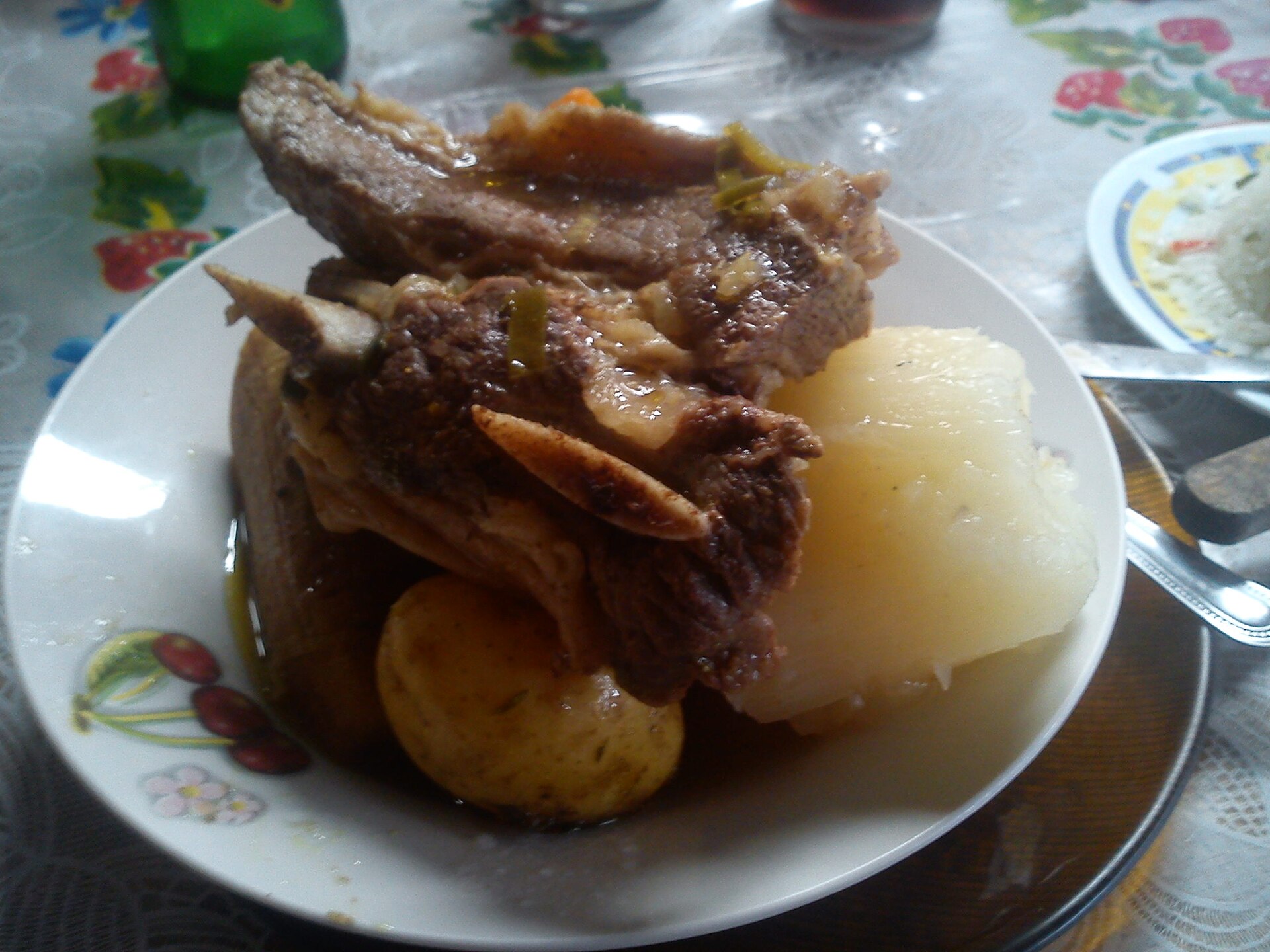 A plate of olla de carne with beef short ribs, a potato, a large piece of yuca and plantain, on a flowered tablecloth