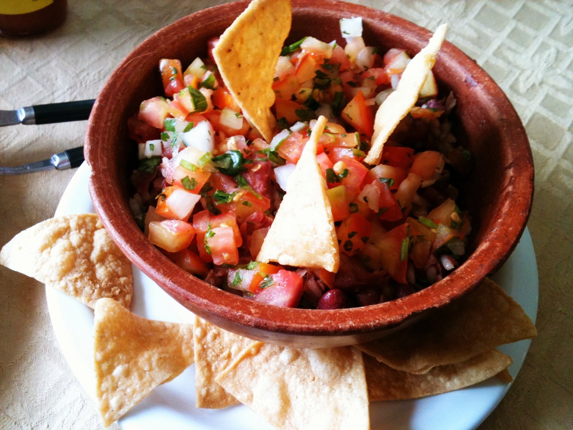 A clay bowl of chifrijo topped with diced tomato, onion and cilantro pico de gallo, with beans visible underneath and tortilla chips around it