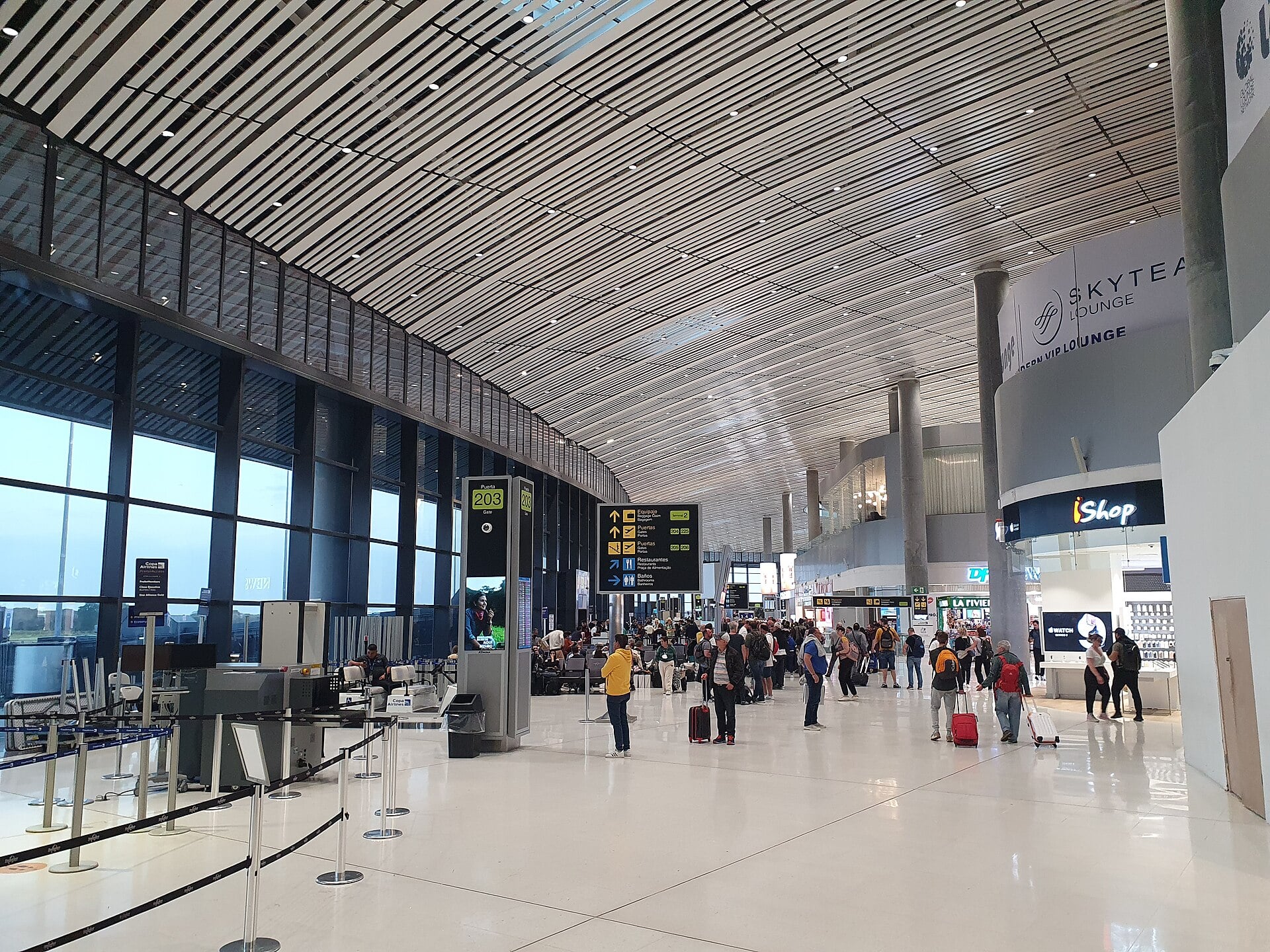 Travellers with roller bags walk through the bright airside hall of Tocumen's Terminal 2 under a slatted ceiling, past a gate sign, shops and a lounge entrance