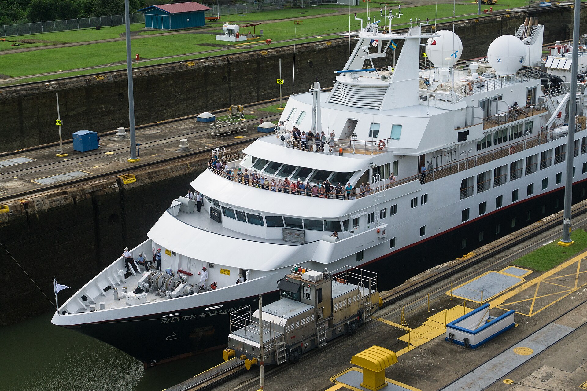 The small white expedition cruise ship Silver Explorer inside a Miraflores Locks chamber, passengers on its decks and a towing locomotive alongside
