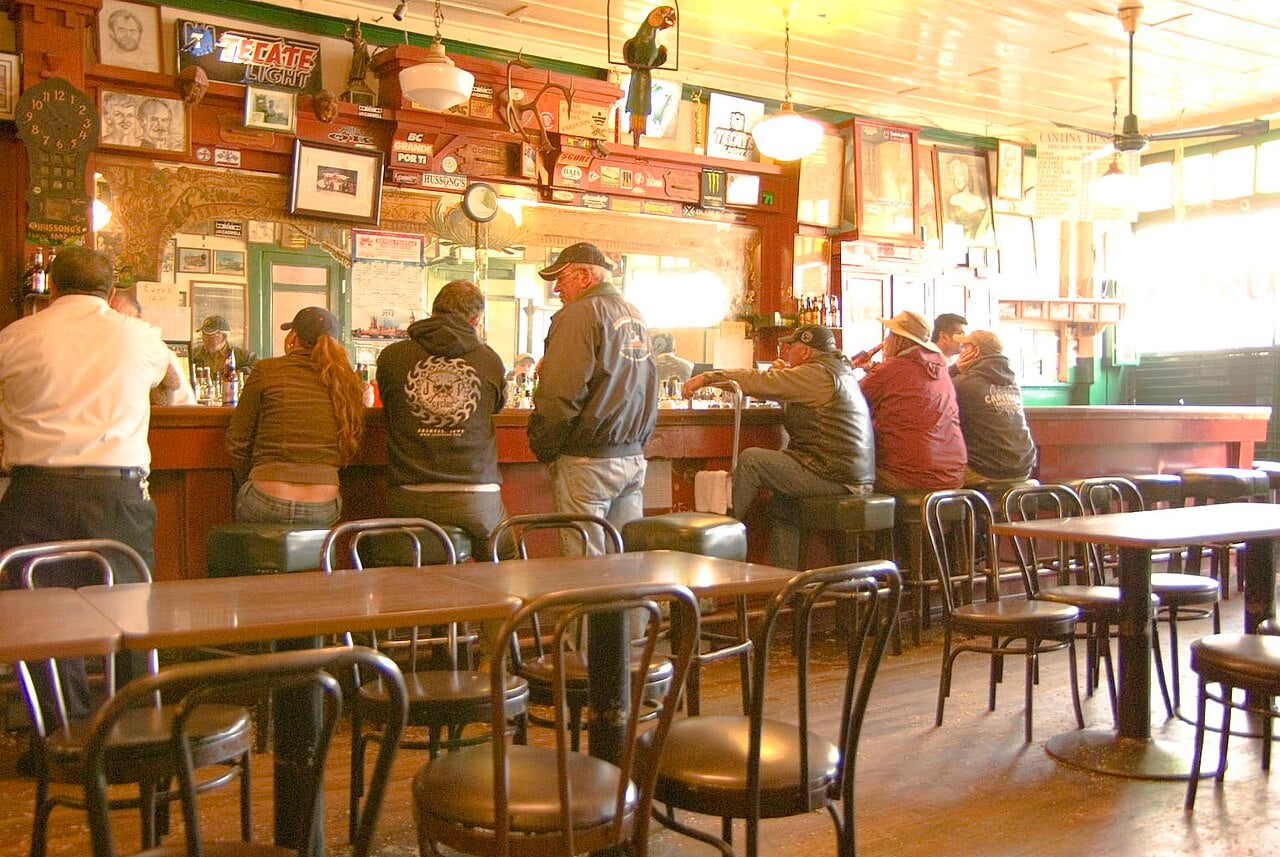 Patrons in jackets and caps stand and sit at the long wooden bar of Hussong's Cantina, with framed photos, a clock and beer signs on the back wall and empty bentwood chairs and tables in front