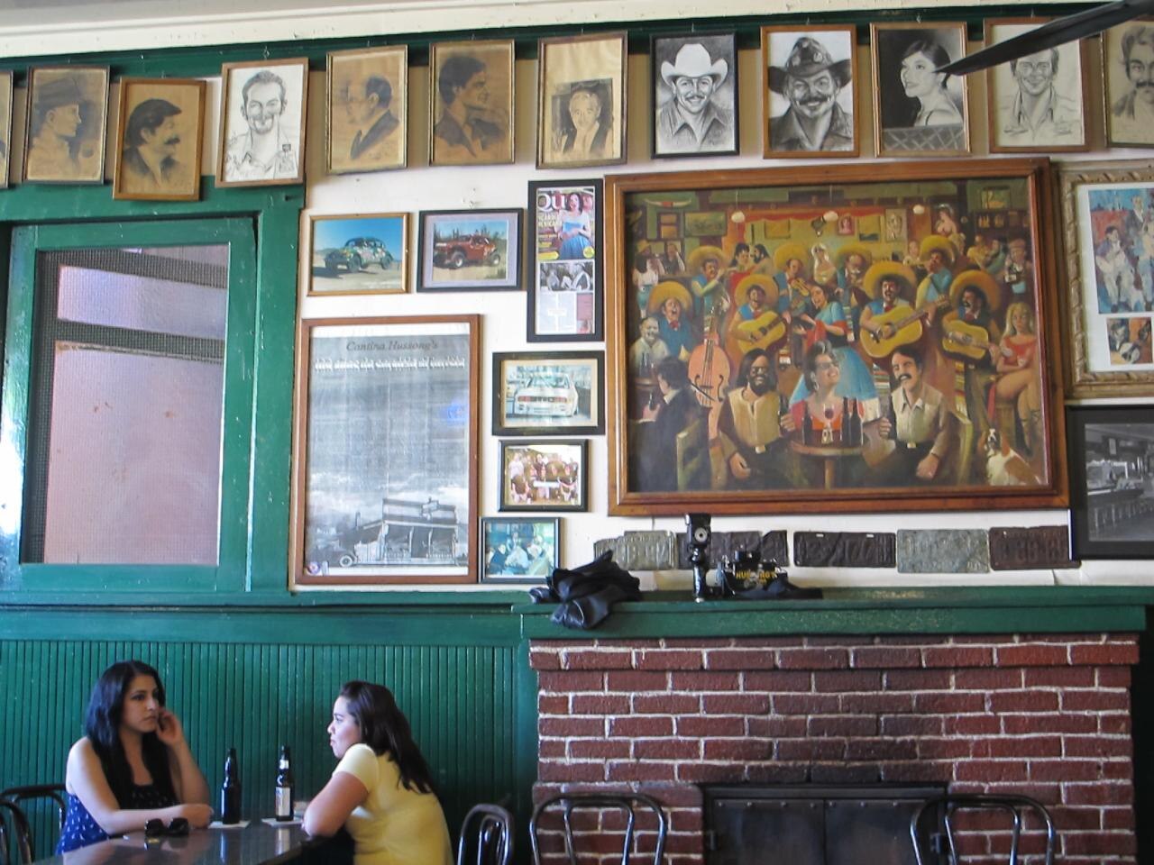 A green-panelled wall at Hussong's Cantina covered in framed portrait drawings, old photos and a large painting of mariachis, above a brick fireplace, with two women talking at a table