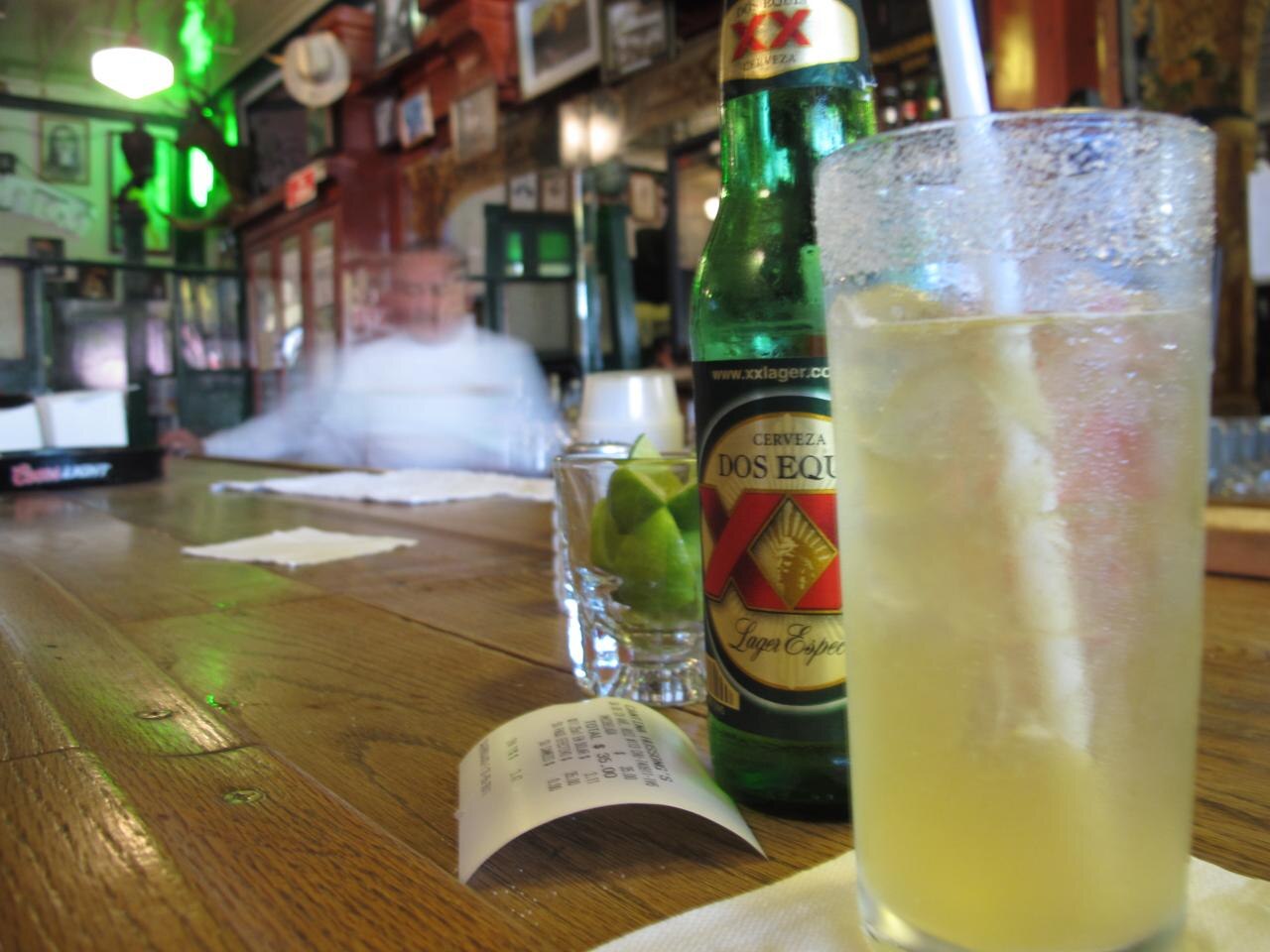 A frosted glass of michelada with a straw, a bottle of Dos Equis lager and a small glass of lime wedges on the worn wooden bar, with a blurred bartender behind