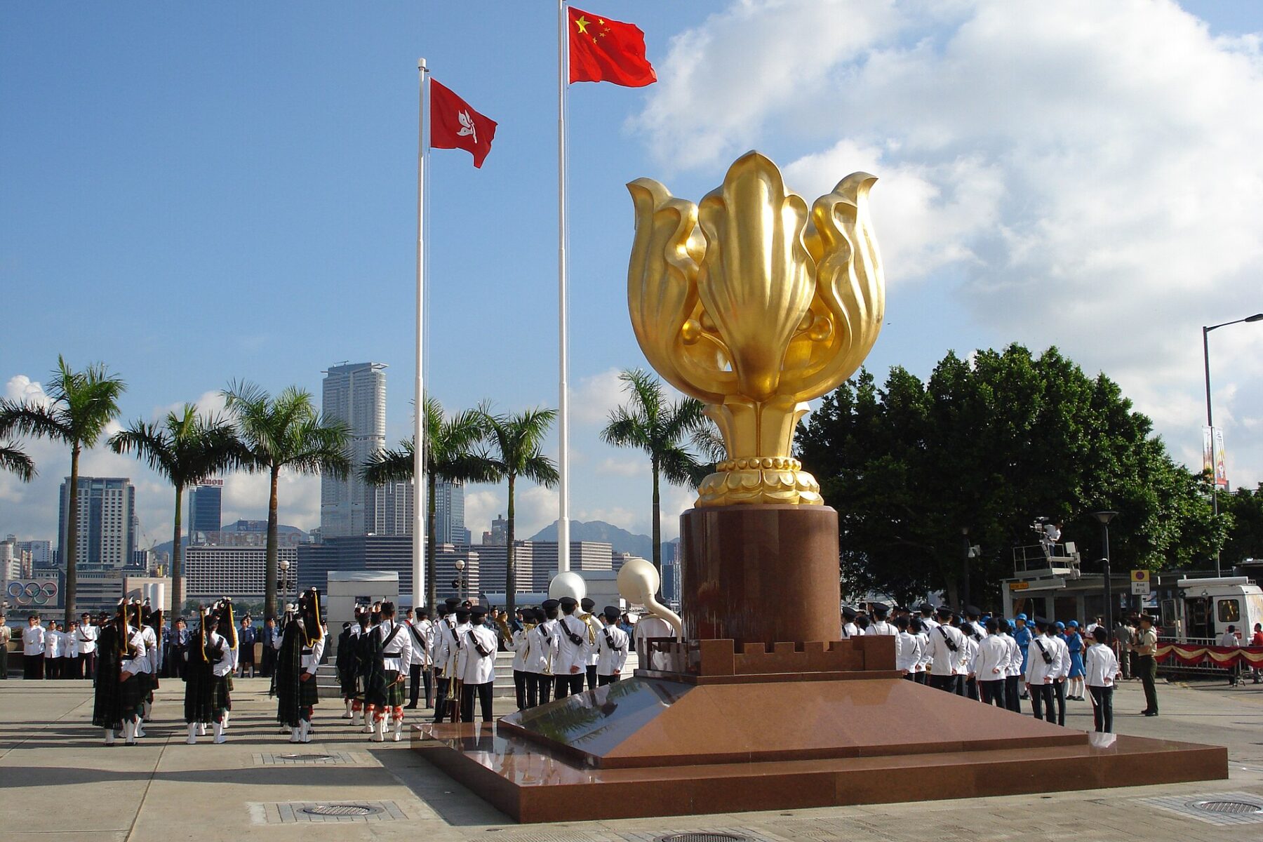 The red flag of the People's Republic of China flies above the red Hong Kong flag with its white bauhinia flower, next to a golden bauhinia sculpture, as a pipe band and uniformed groups stand in formation