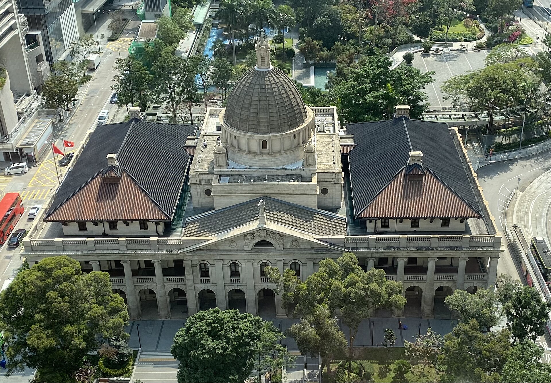 Aerial view of a domed neoclassical granite building with two dark pitched roofs and a colonnaded front, surrounded by trees and roads in Central, Hong Kong