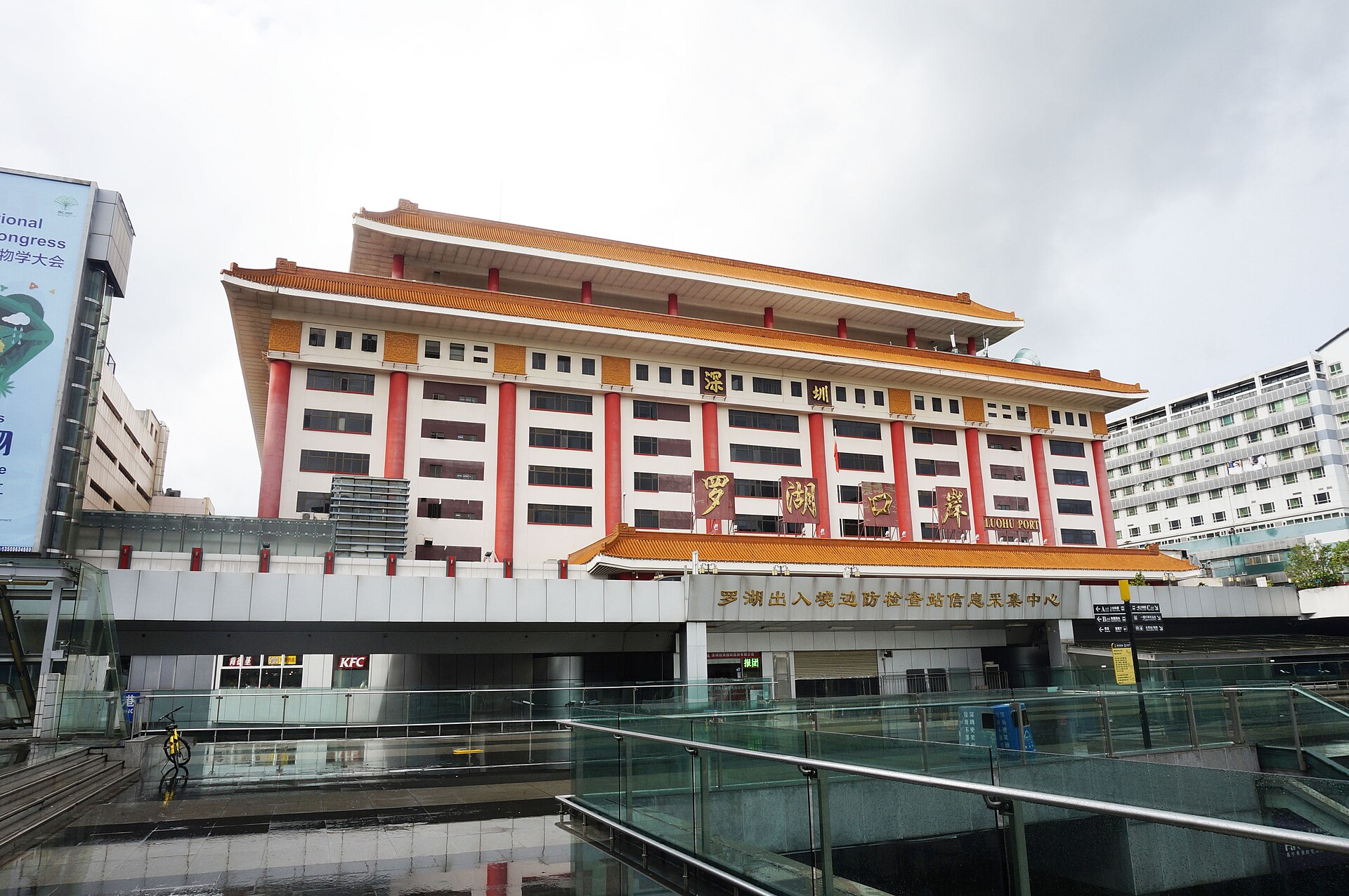 A large white building with red pillars and an orange tiled roof, bearing the Chinese characters for Shenzhen Luohu Port, seen across a glass-sided walkway on a cloudy day