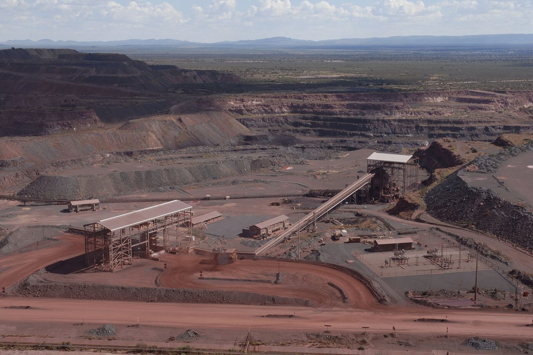 Terraced red walls of a vast open-pit iron ore mine with conveyor structures, sheds and haul roads in the foreground under a cloudy sky