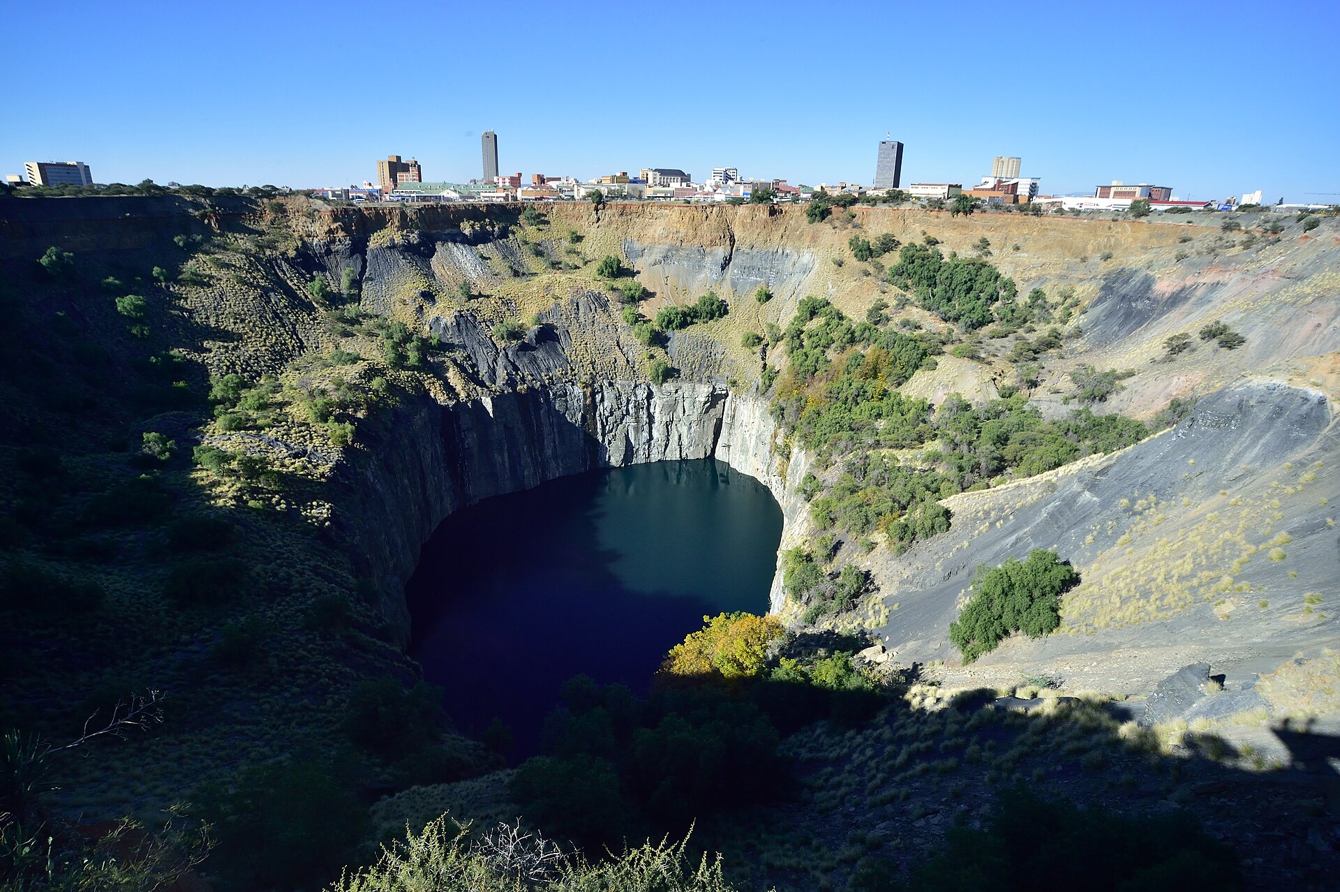 A vast open pit with steep rock walls and dark water at the bottom, with the low skyline of Kimberley on the far rim under a clear sky